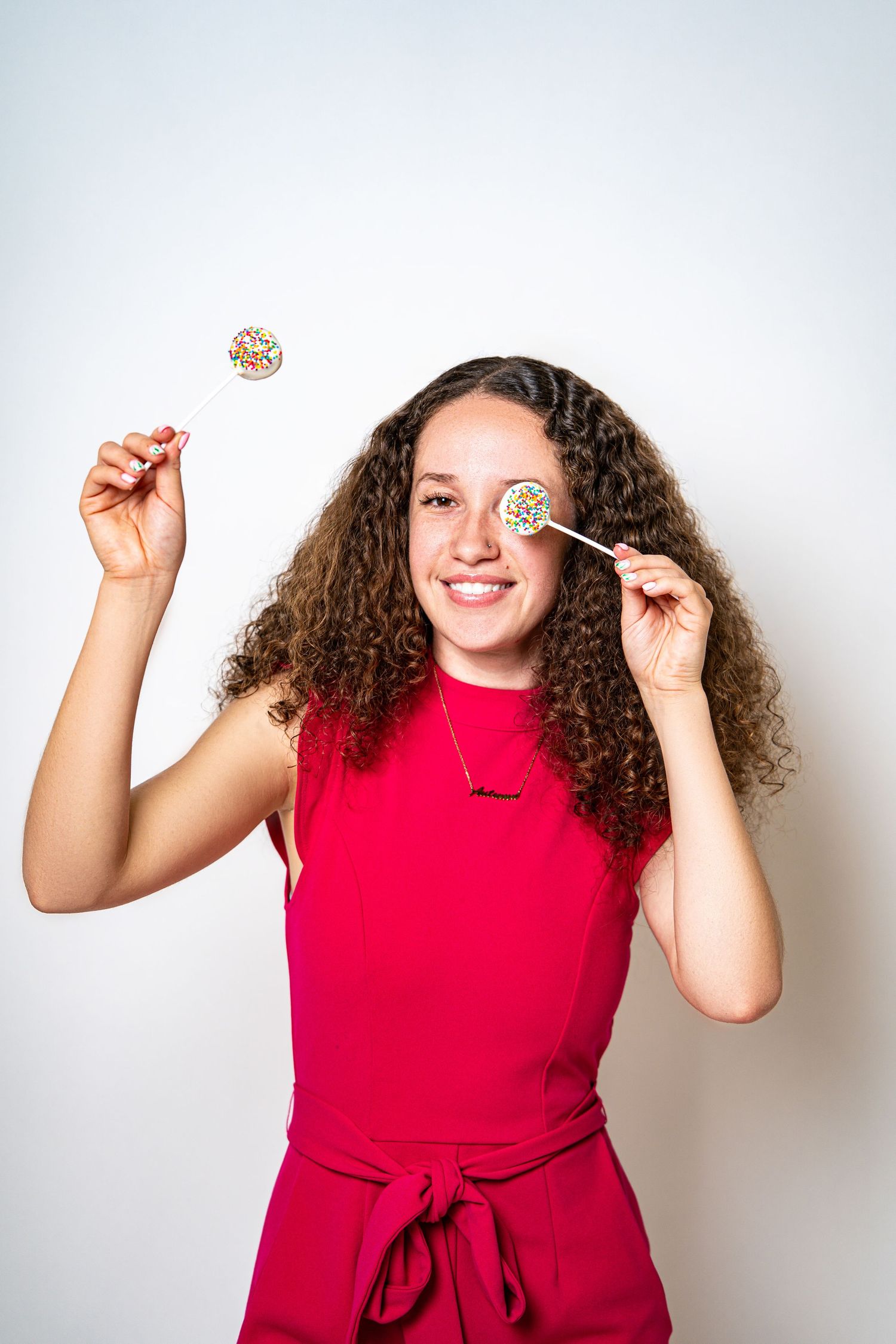 Happy young woman with curly hair holding two colorful sprinkle cake pops over her eyes in red dress.