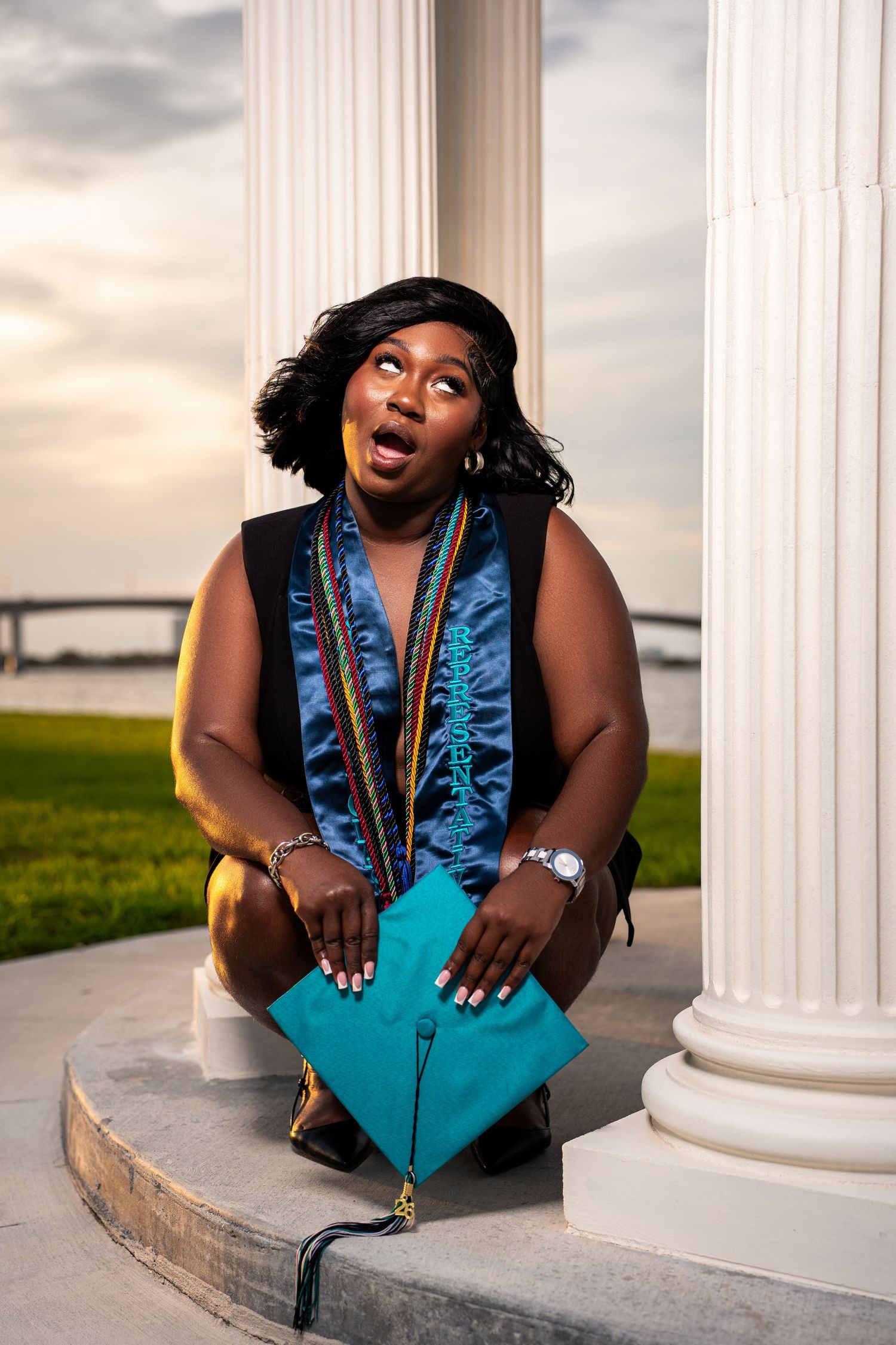 Black female graduate crouching beside white column holding teal cap with surprised expression at sunset.