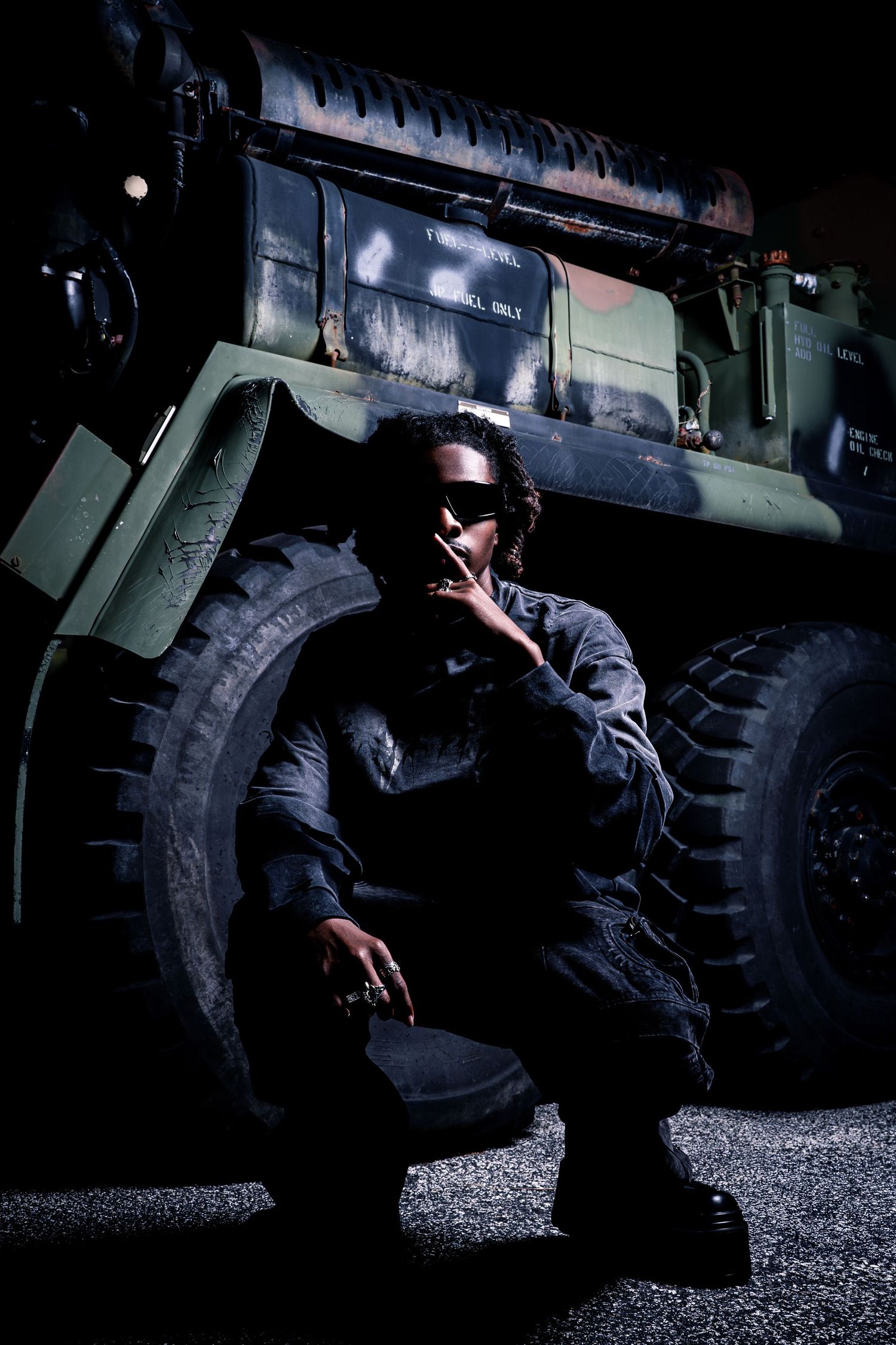A person in dark military-style clothing crouches beside a large military truck in dramatic low lighting.