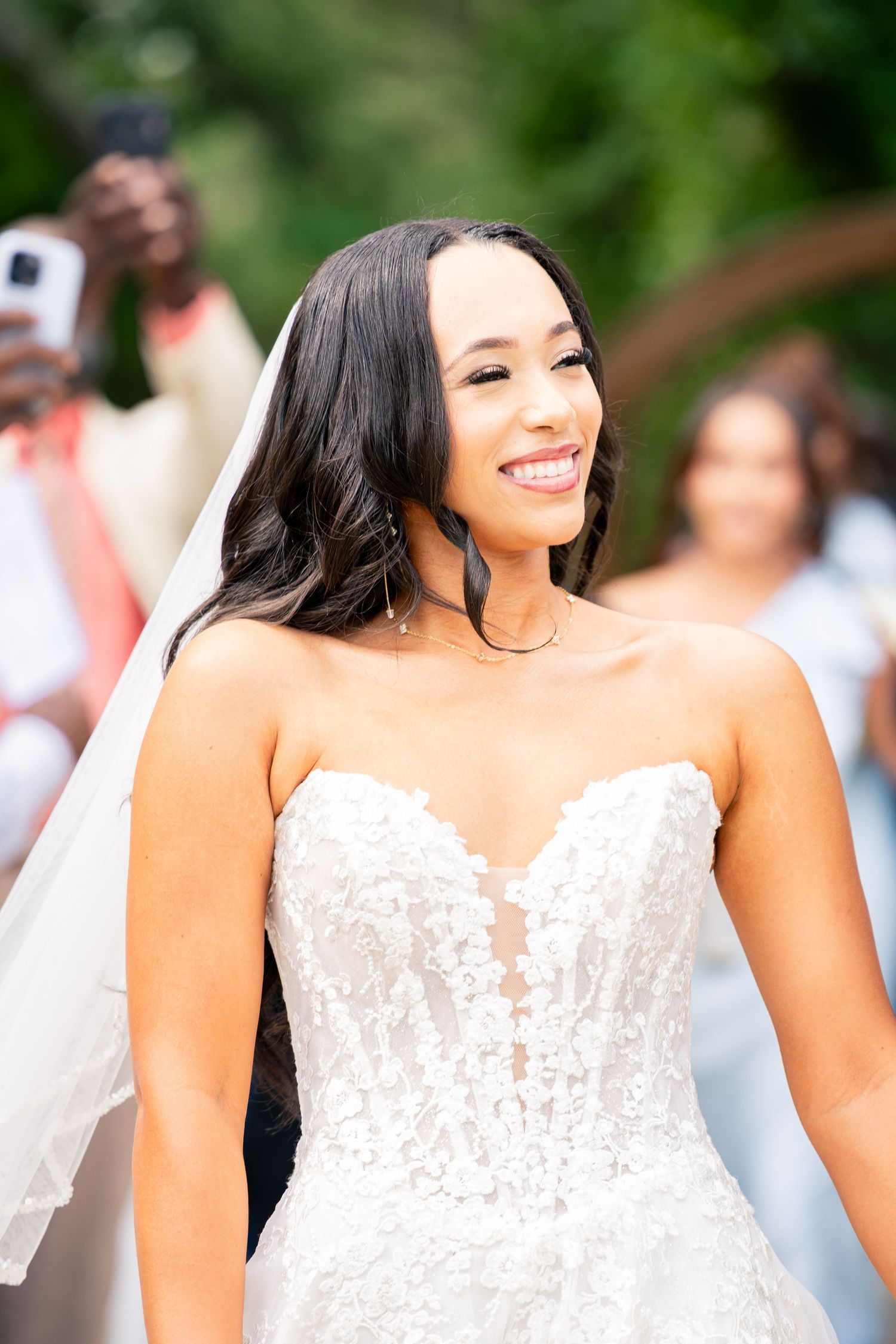 Smiling bride in strapless lace wedding gown and veil walks down the aisle at outdoor ceremony.