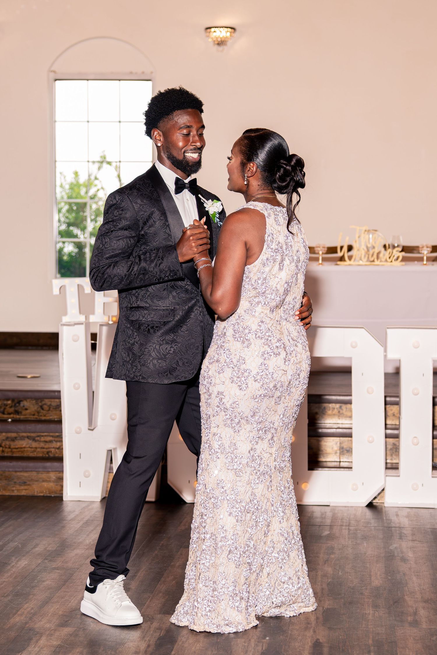 Smiling groom in black tuxedo dances with woman in silver sequin gown at wedding reception.