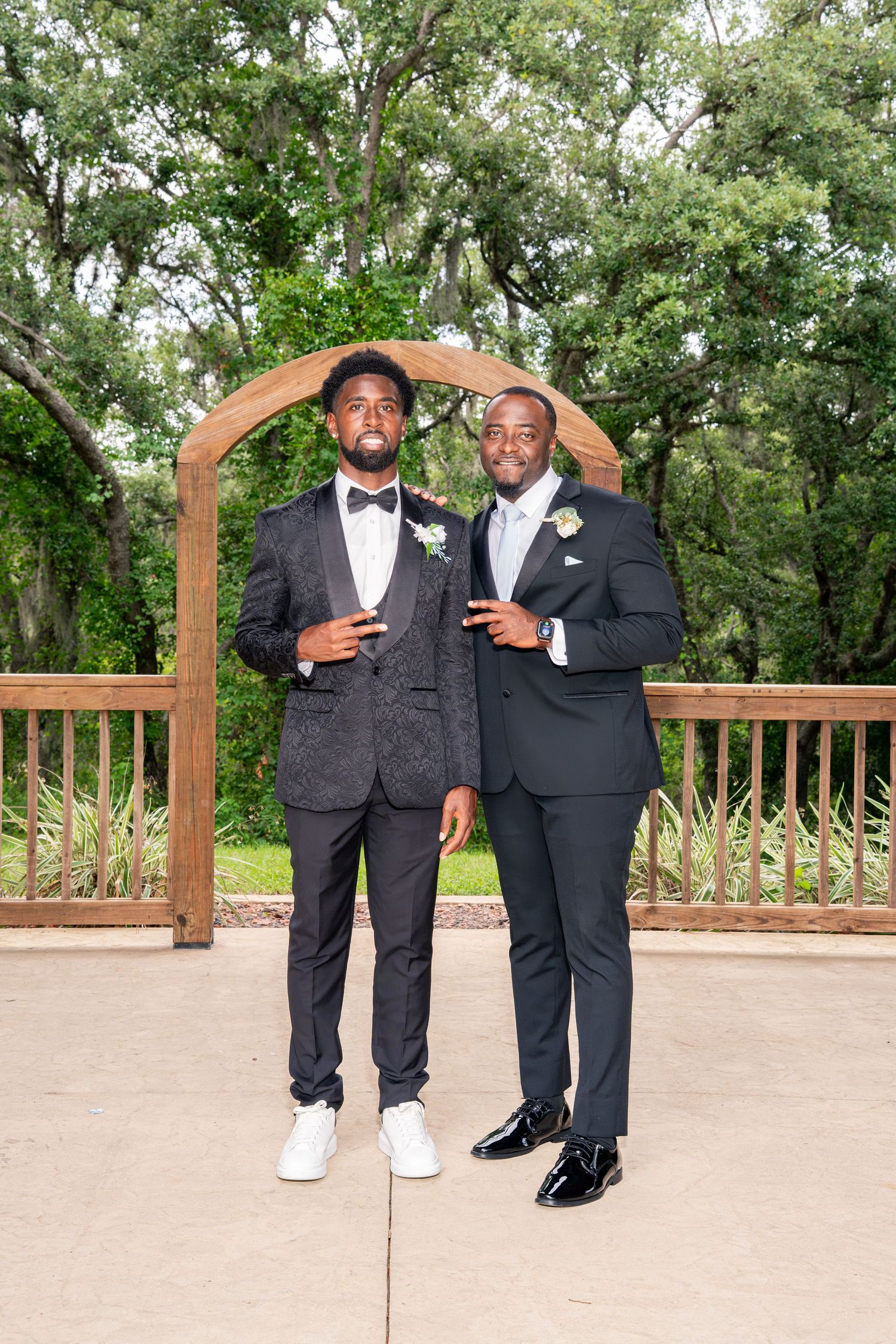 Two well-dressed groomsmen pose confidently in black tuxedos under wooden arch at outdoor wedding venue.