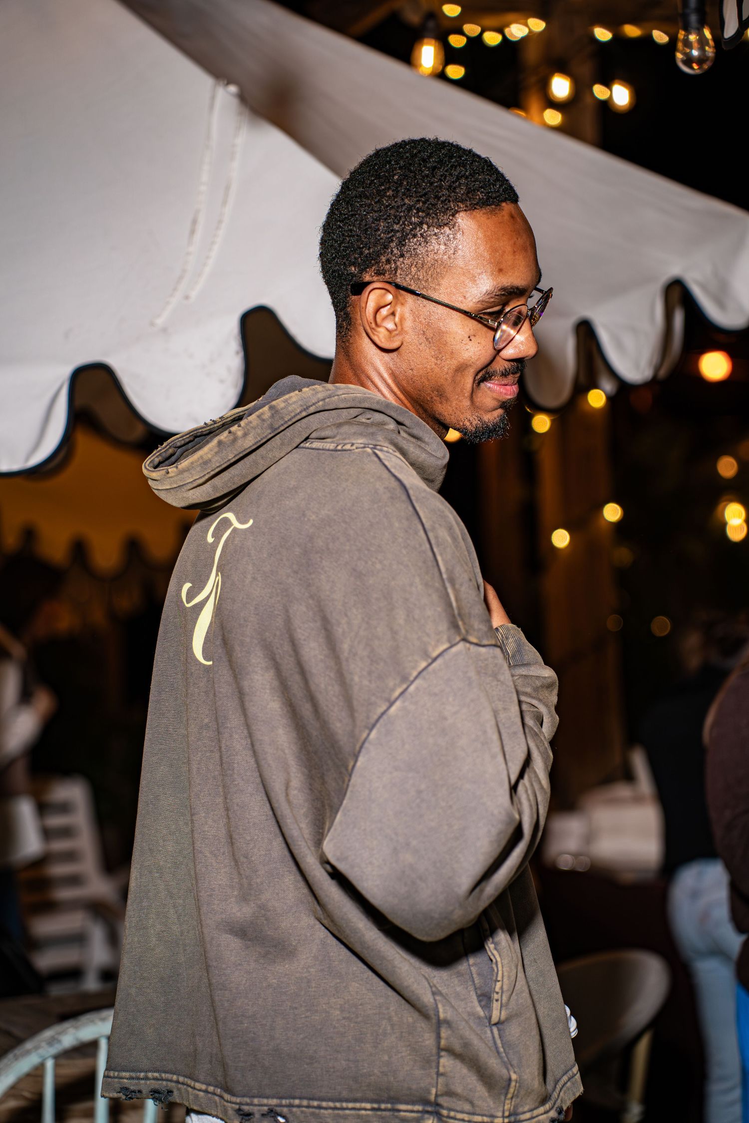 Smiling man in olive hoodie with gold monogram detail at outdoor evening gathering.