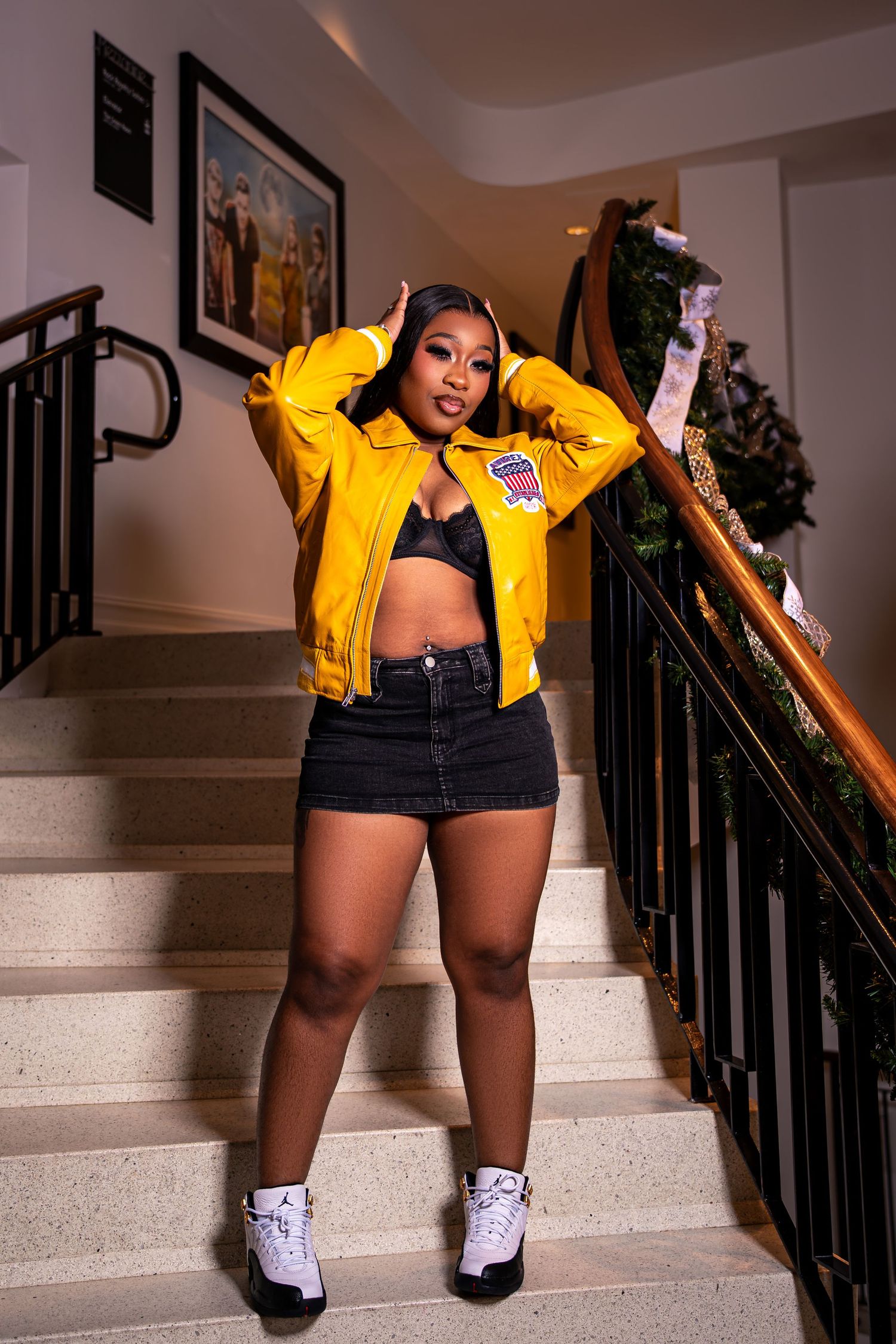 Confident Black woman in yellow varsity jacket and black mini skirt poses on carpeted staircase.