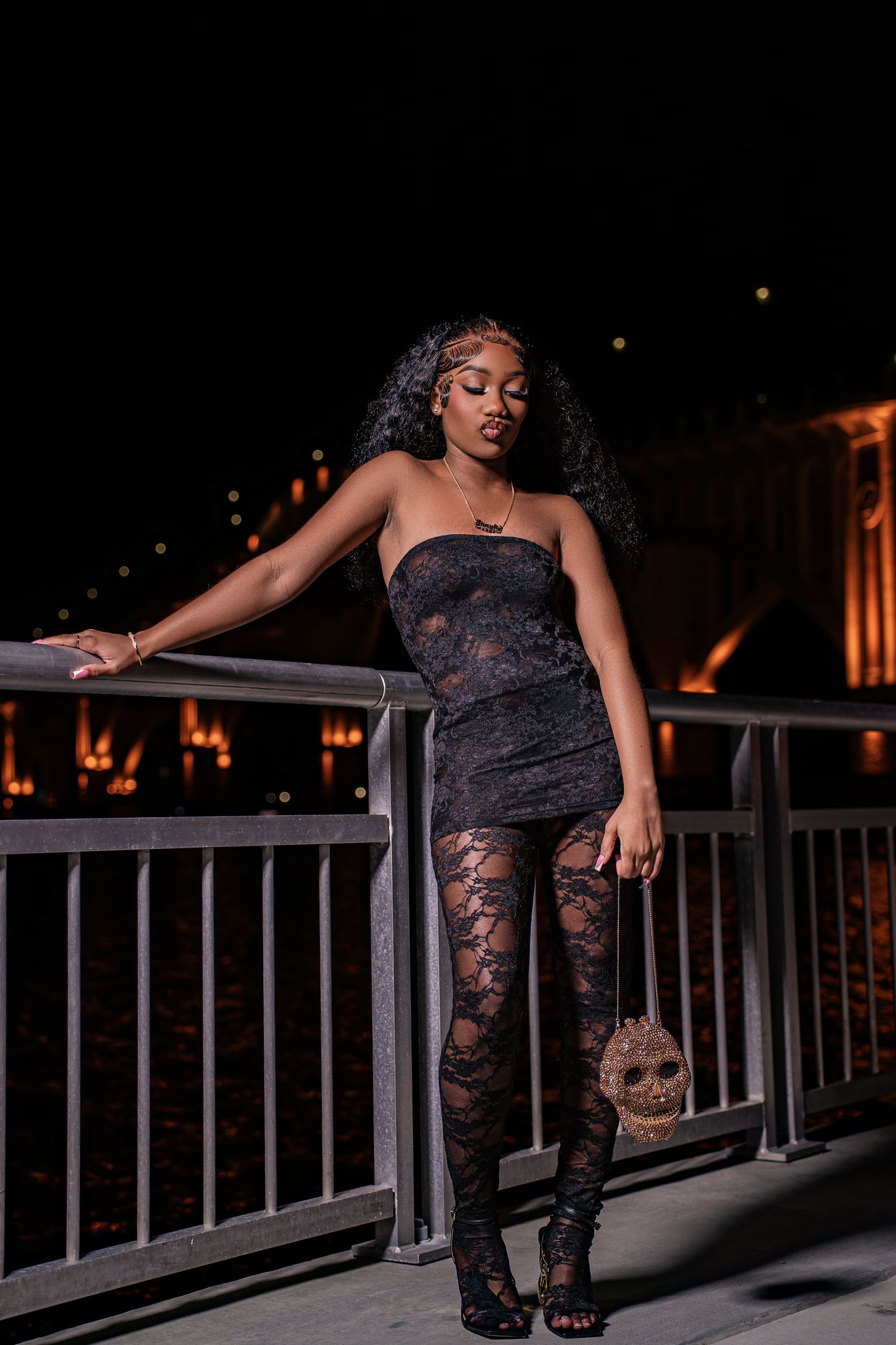 Elegant woman in black lace strapless mini dress and tights posing on bridge railing at night holding decorative mask.
