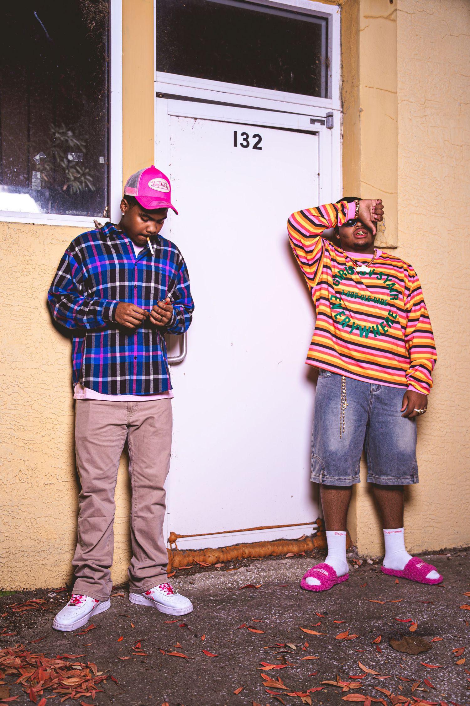 Two young men in bold streetwear standing by white door 132 on yellow building wearing plaid shirt and striped sweater.