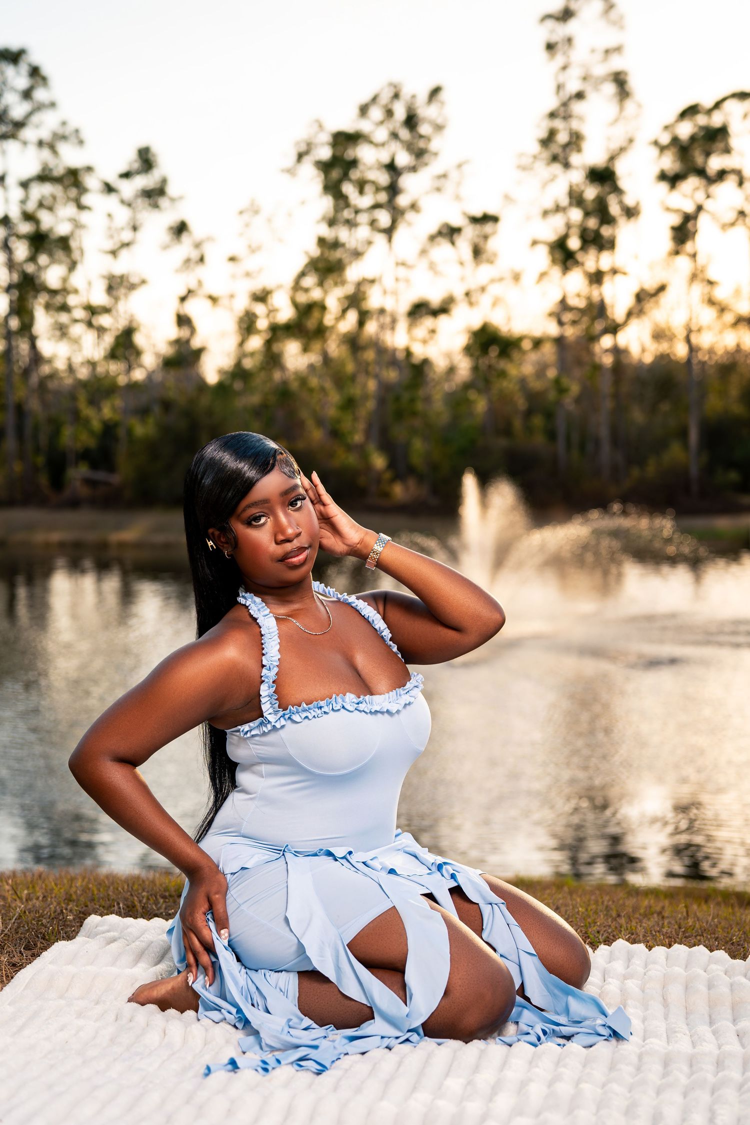 Black woman in light blue fringe dress poses gracefully on white blanket near sunset pond.