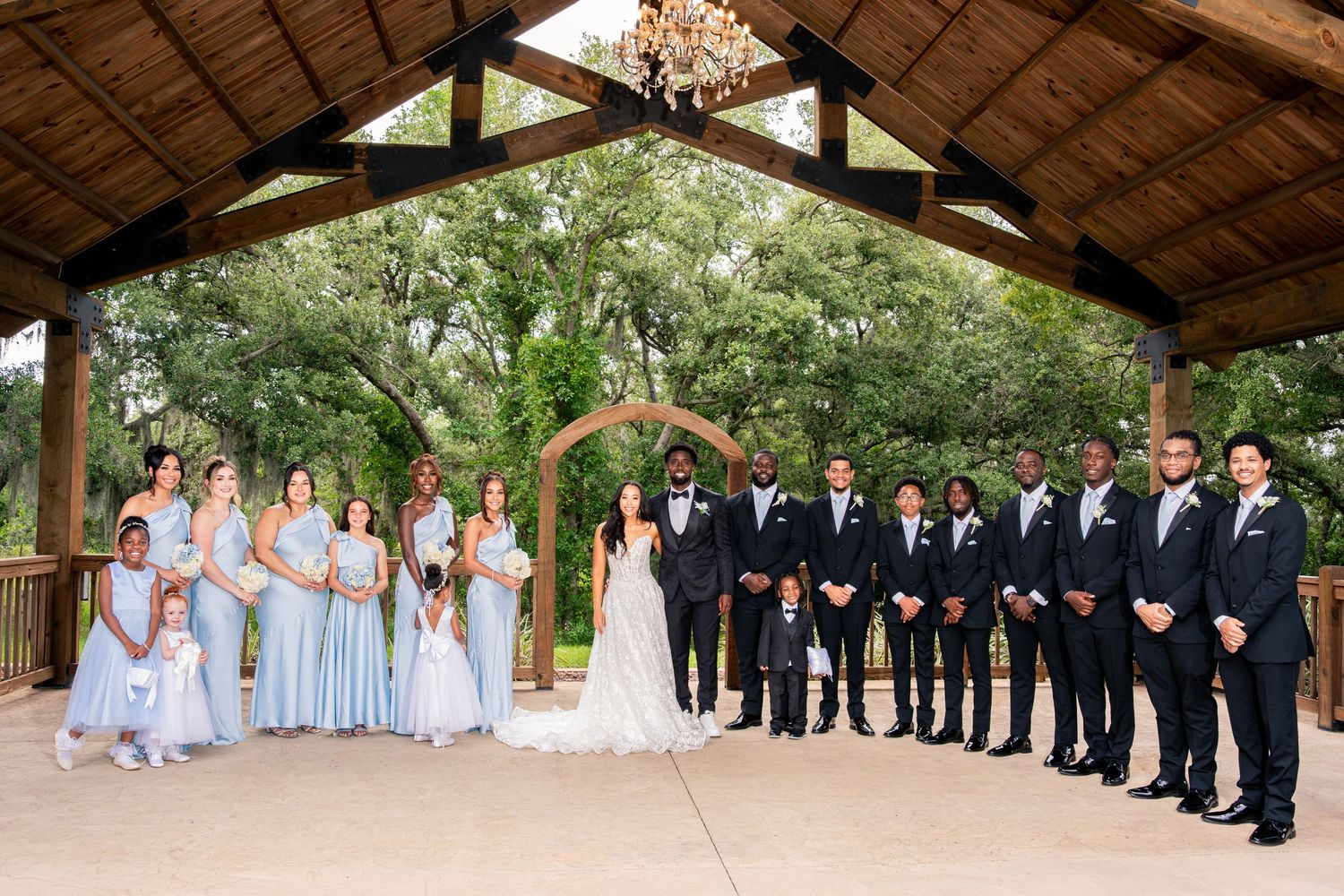 Wedding party poses under wooden pavilion with bridesmaids in blue gowns and groomsmen in black suits.