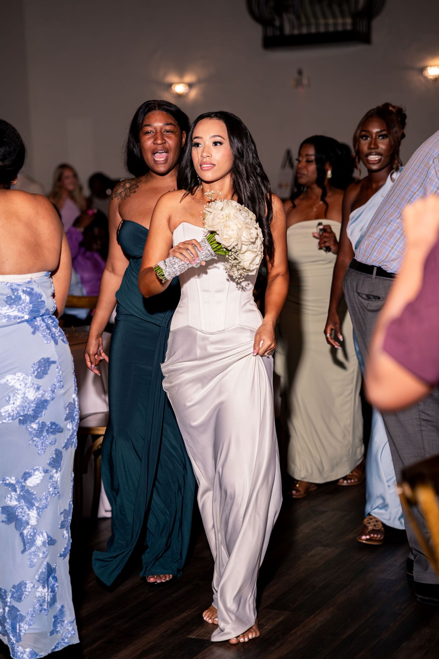 Bride in white jumpsuit dancing with guests on crowded reception dance floor holding bouquet.