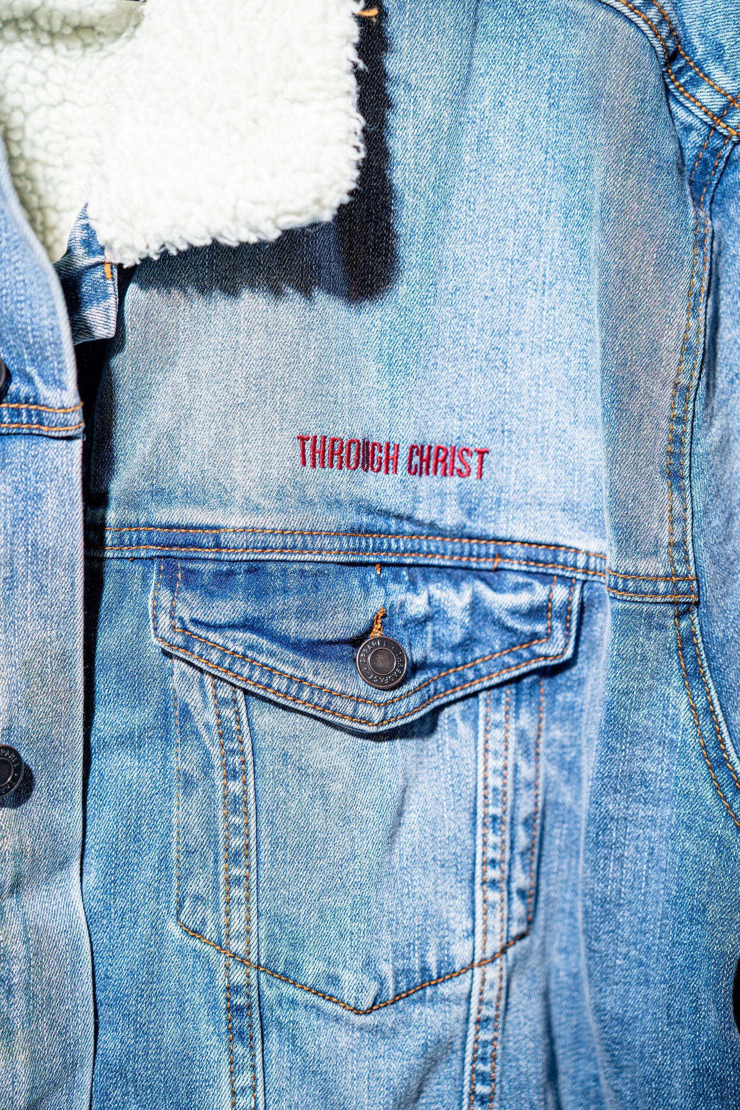 Denim sherpa jacket with red embroidered Through Christ text on chest pocket.