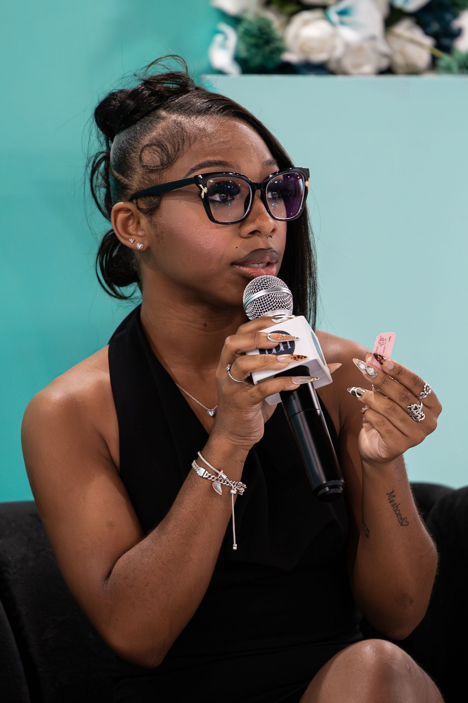 Young woman in black dress speaks into microphone at DDH Entrepreneur Success conference, wearing glasses and bracelets.