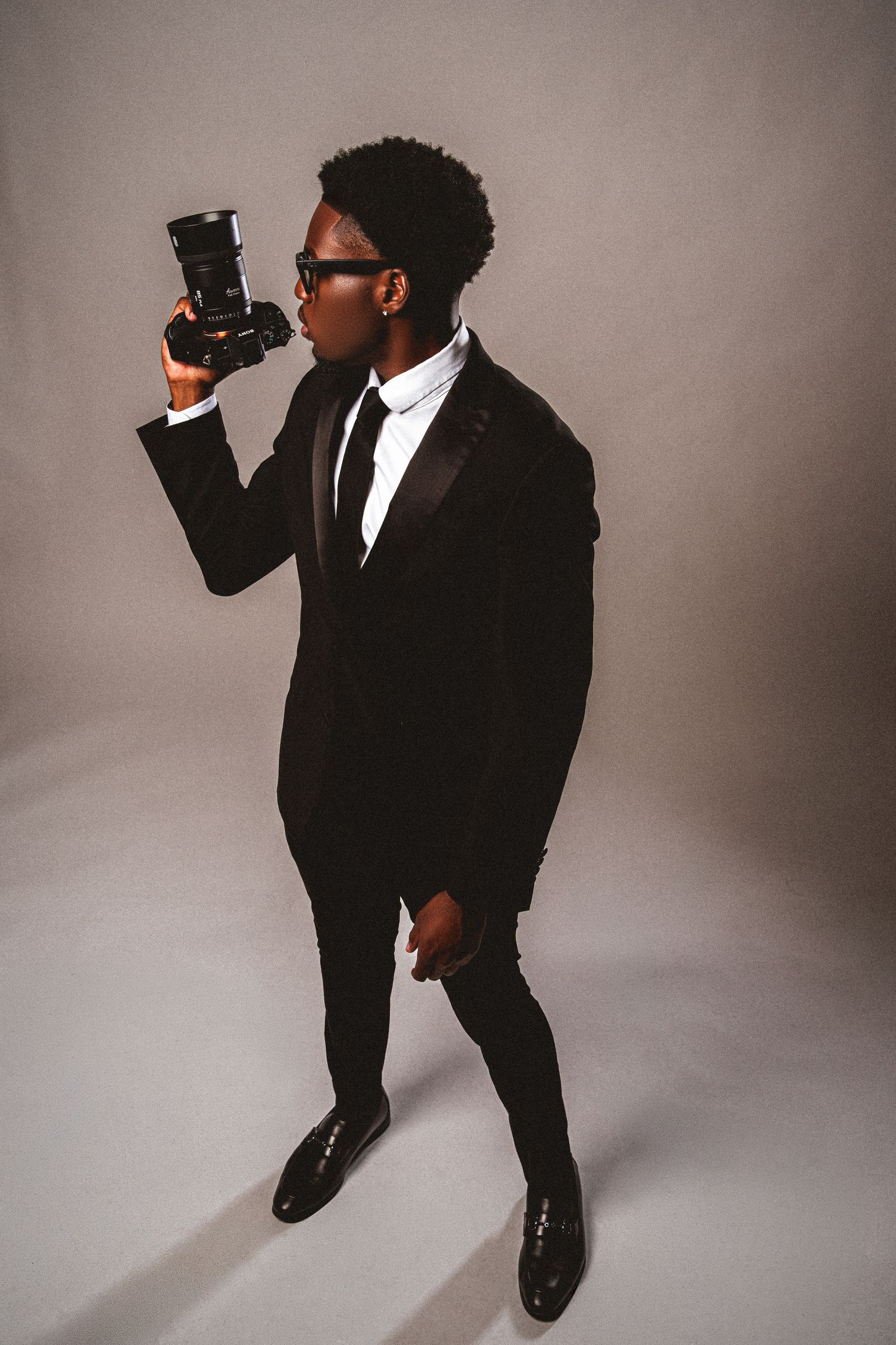 Stylish man in black tuxedo holding a camera, wearing sunglasses, posing against a gray studio background.
