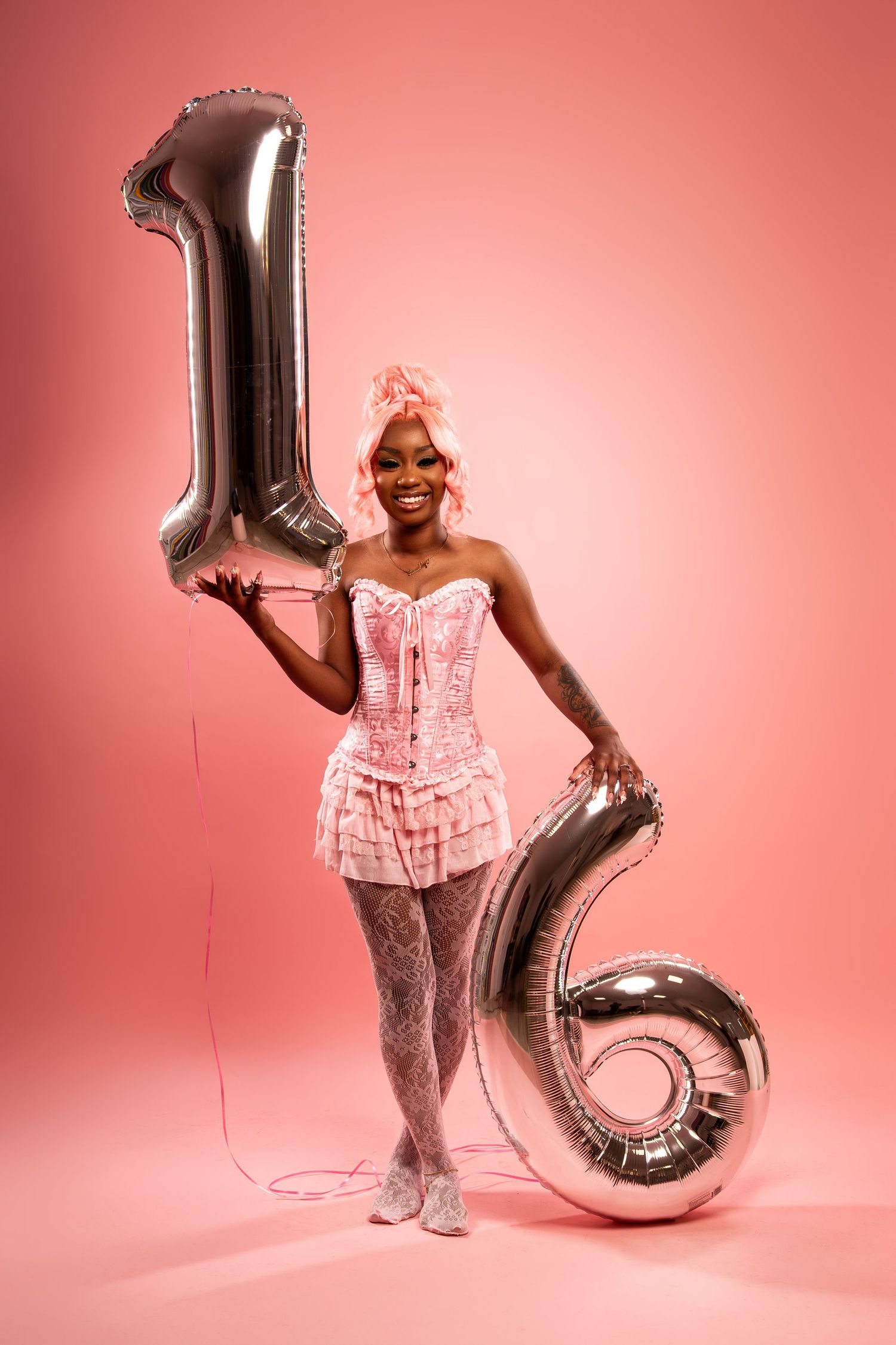 Woman in pink corset outfit holding silver number 16 balloons on pink background.
