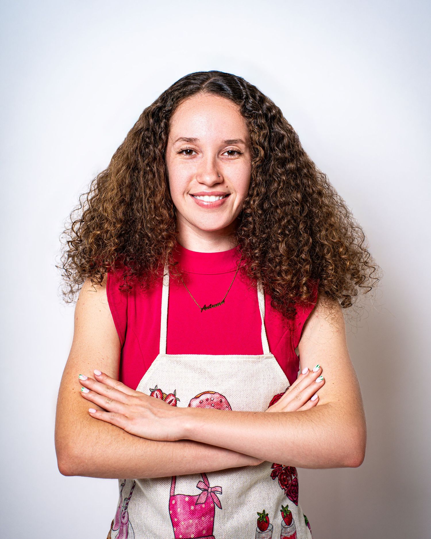 Smiling young woman with curly hair wearing a red top and decorative baking apron with arms crossed.