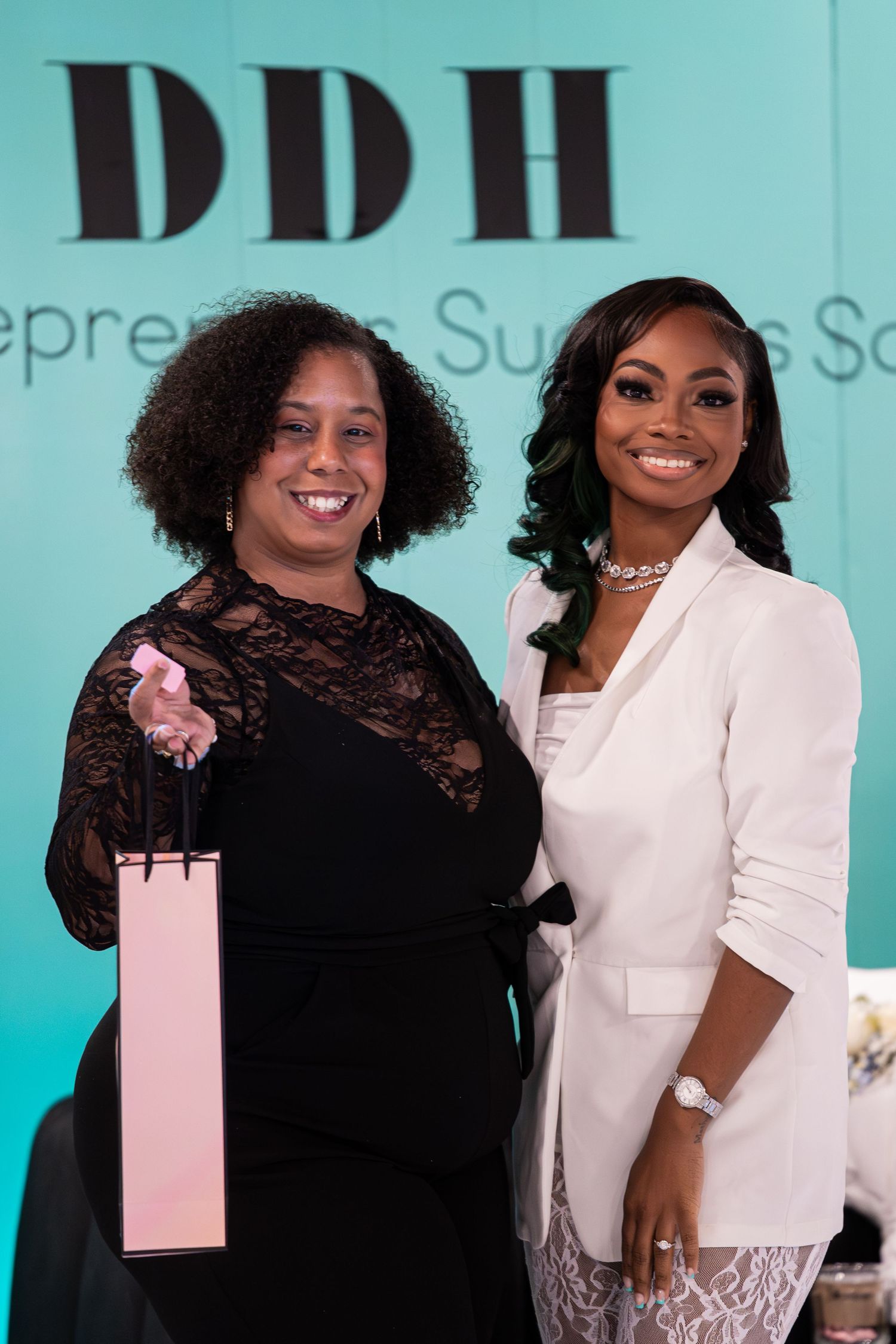 Two women smile together at DDH Entrepreneurial Success Summit, one holding pink gift bag.