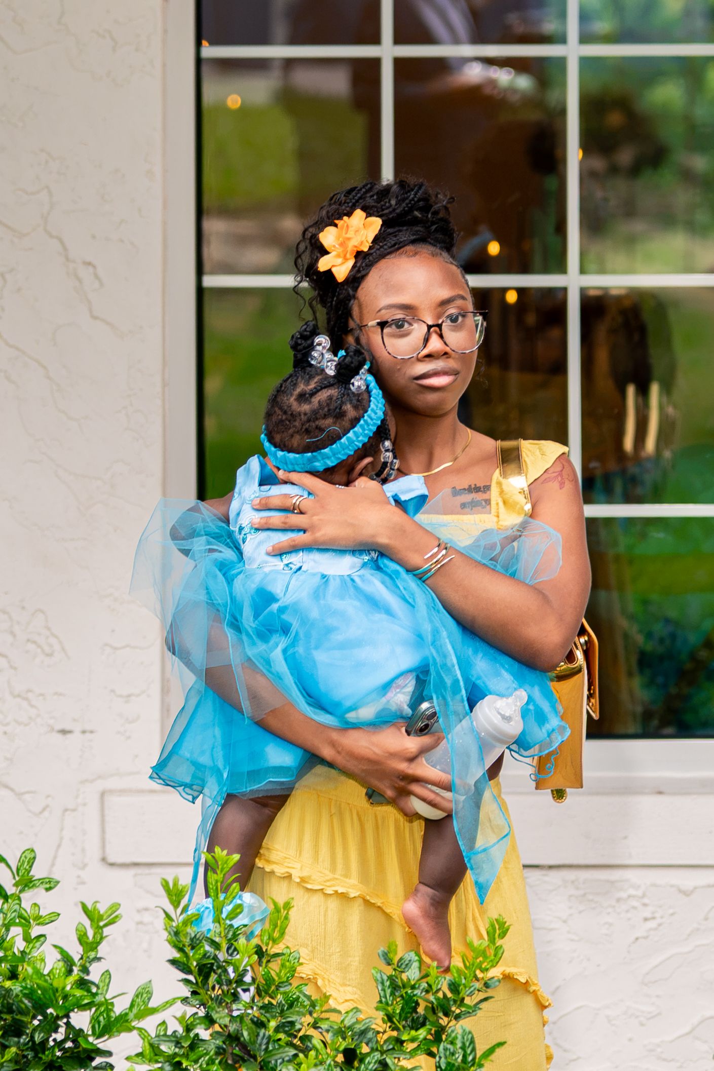 Young woman in yellow dress holds baby in blue tutu outfit outdoors.