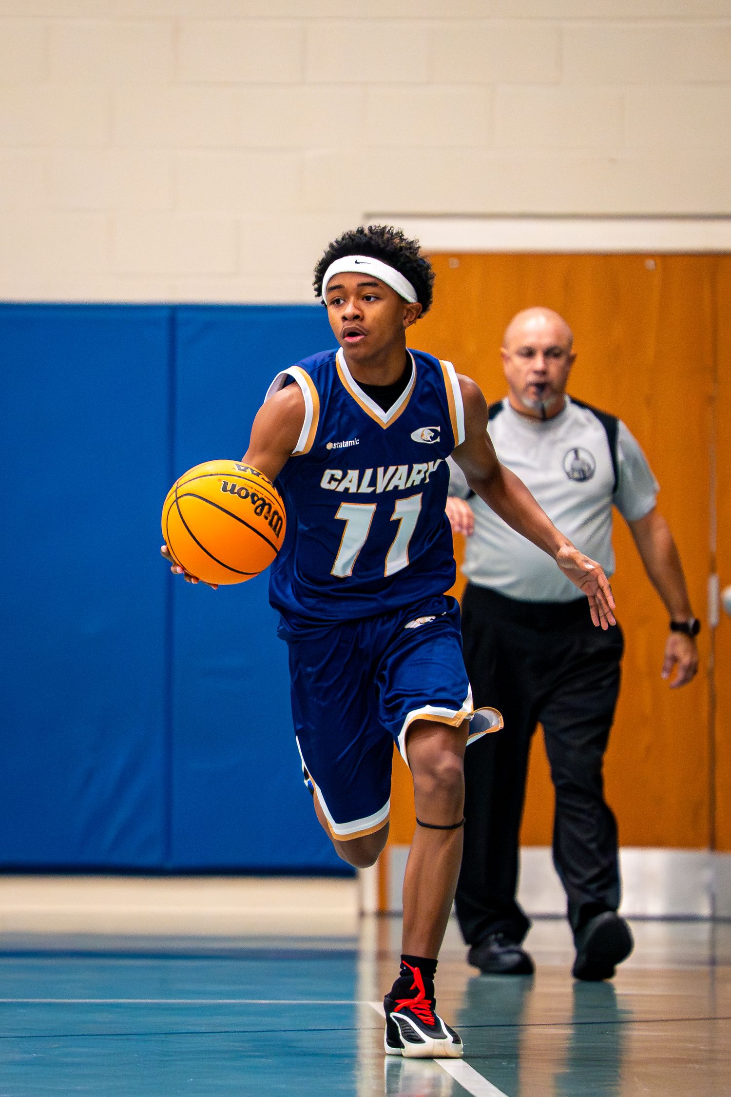 Calvary basketball player #11 dribbles ball upcourt in blue uniform with referee following behind.
