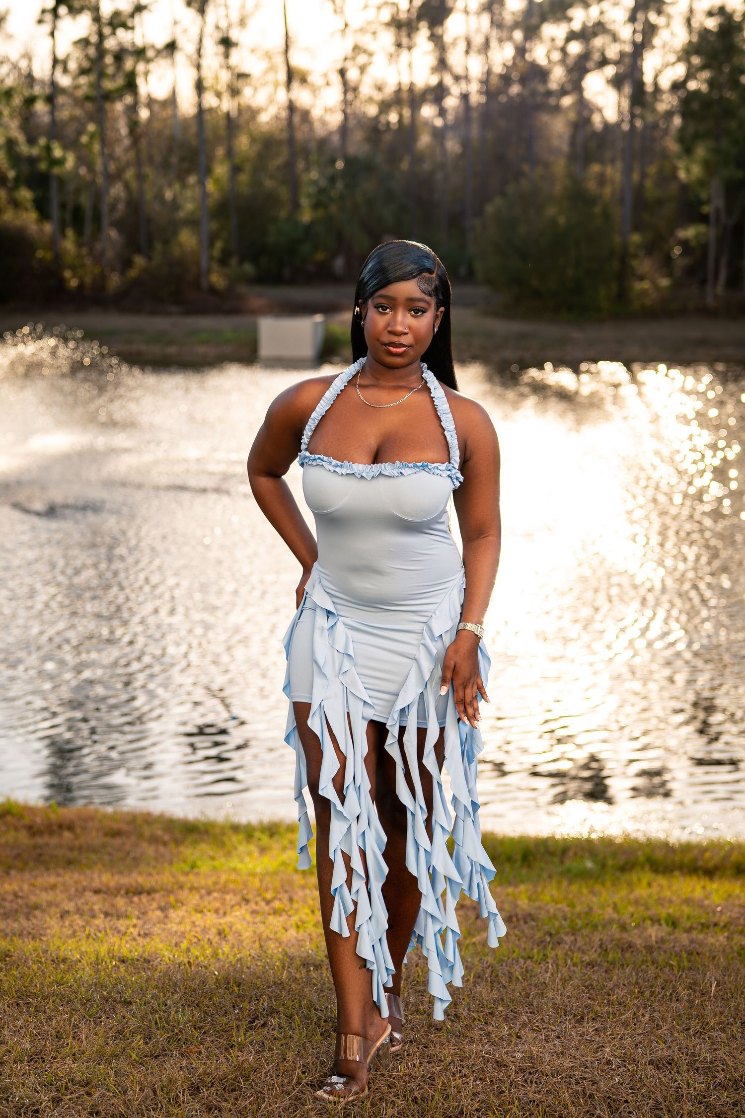 Black woman in light blue fringe halter dress stands confidently by shimmering lake at dusk.