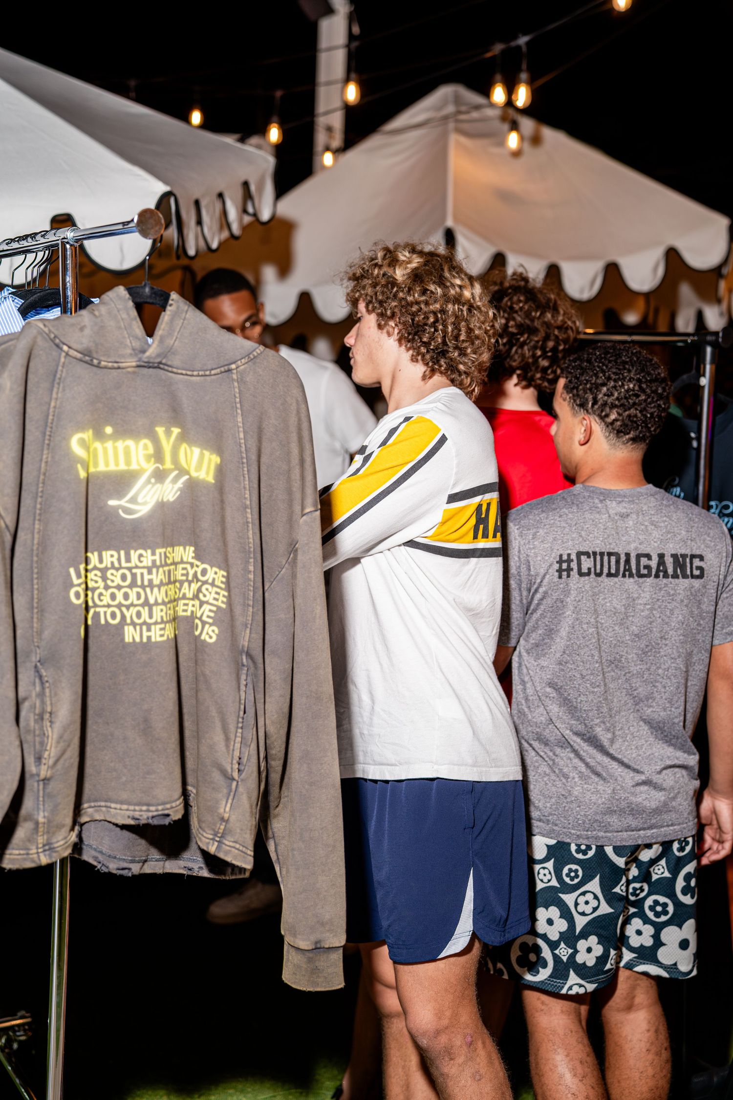 Young men browsing Shine Your Light Christian apparel on rack at outdoor night event.