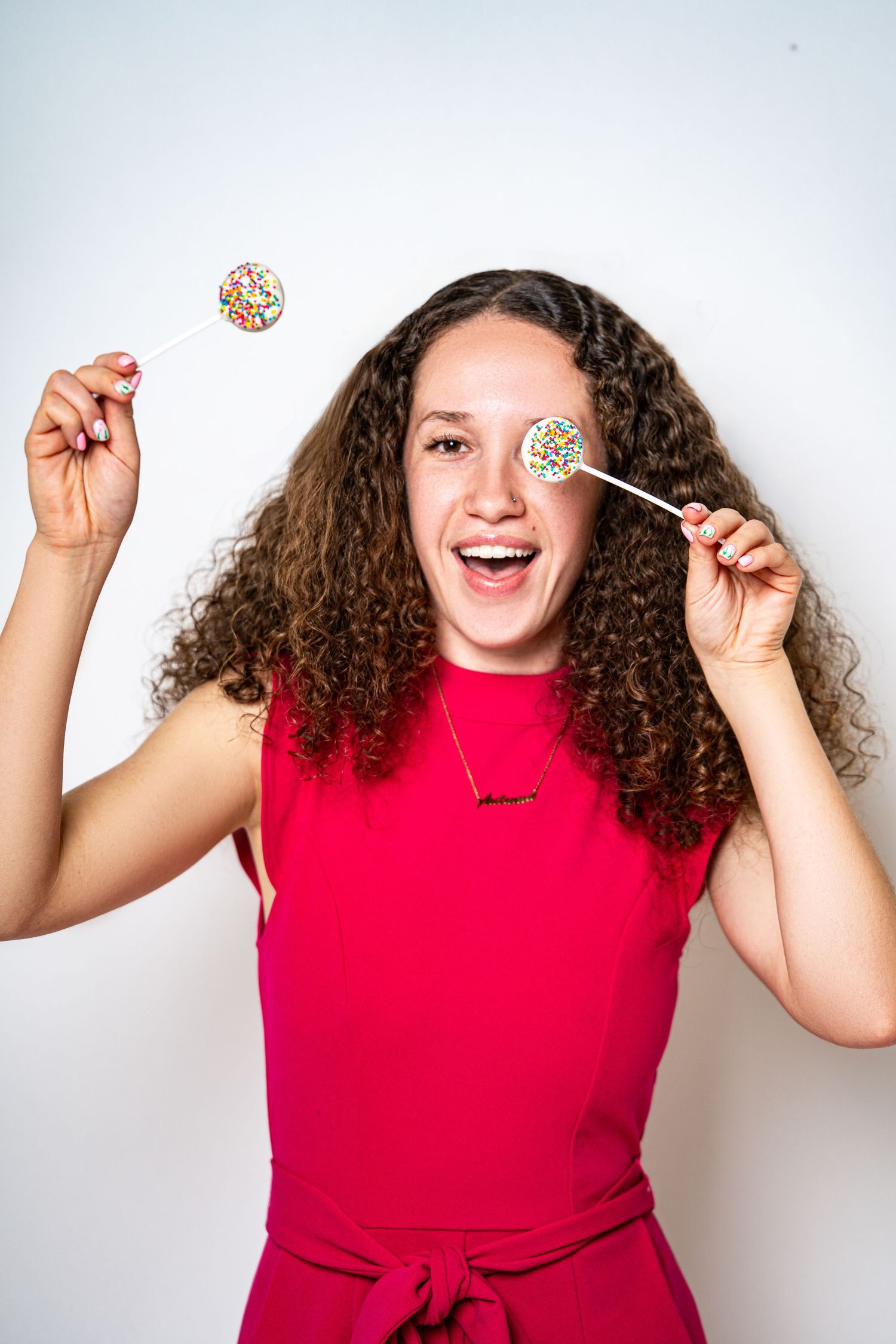 Happy curly-haired woman in red dress playfully holding two colorful sprinkle lollipops over her eyes.