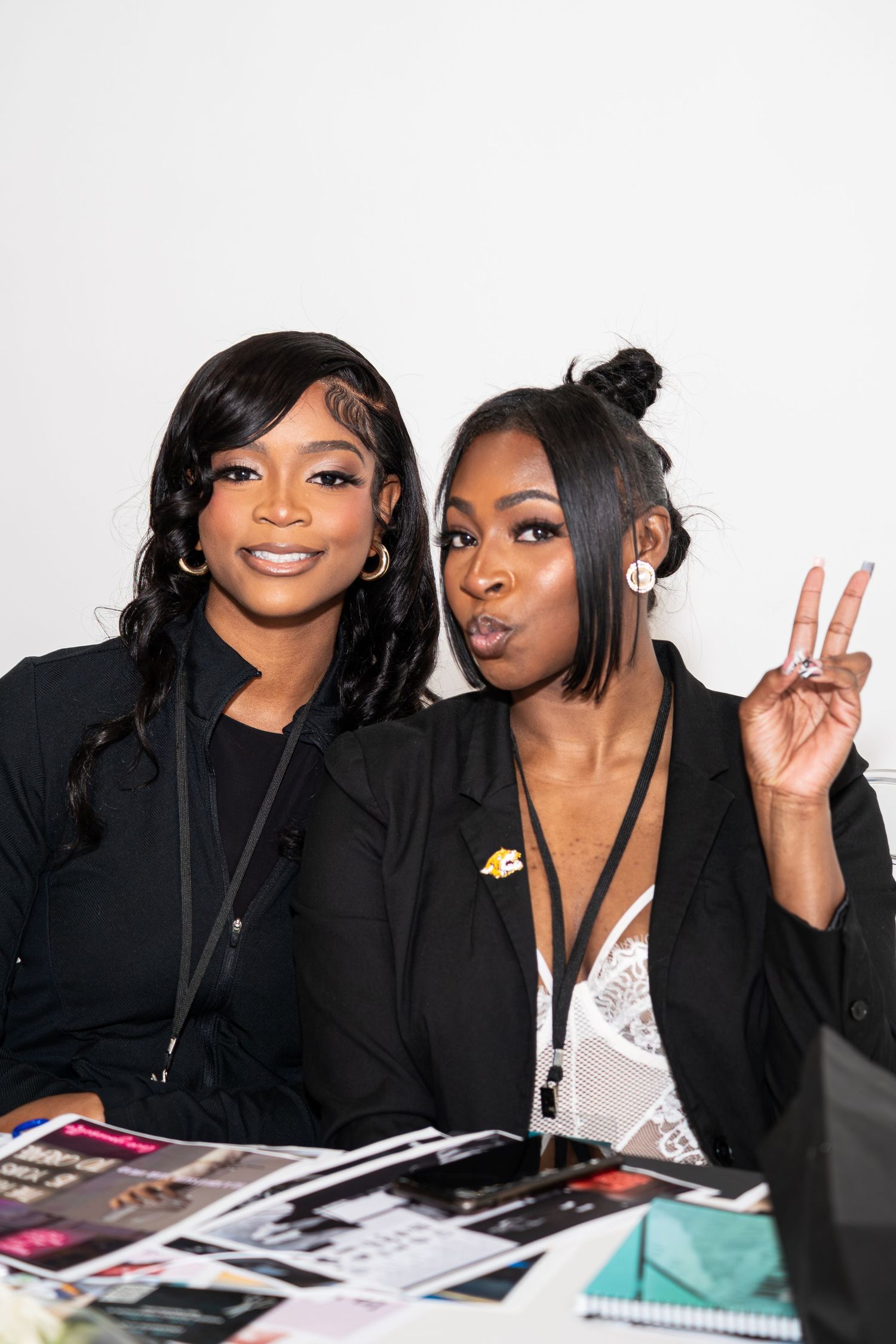 Two young women smile and pose at a table covered with magazines at a networking event.