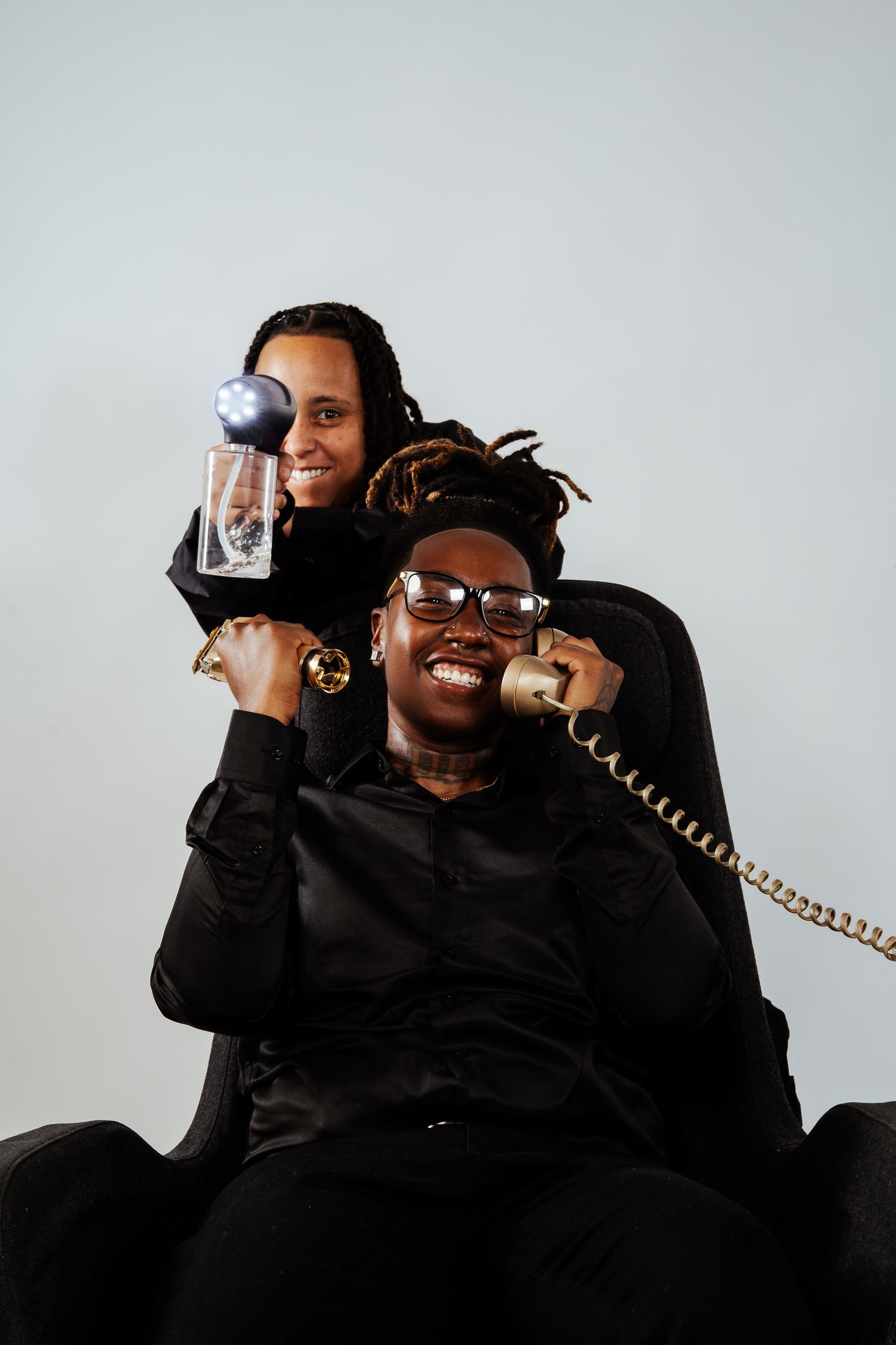 Two people in black outfits posing with vintage phone and illuminated glass cup in studio.