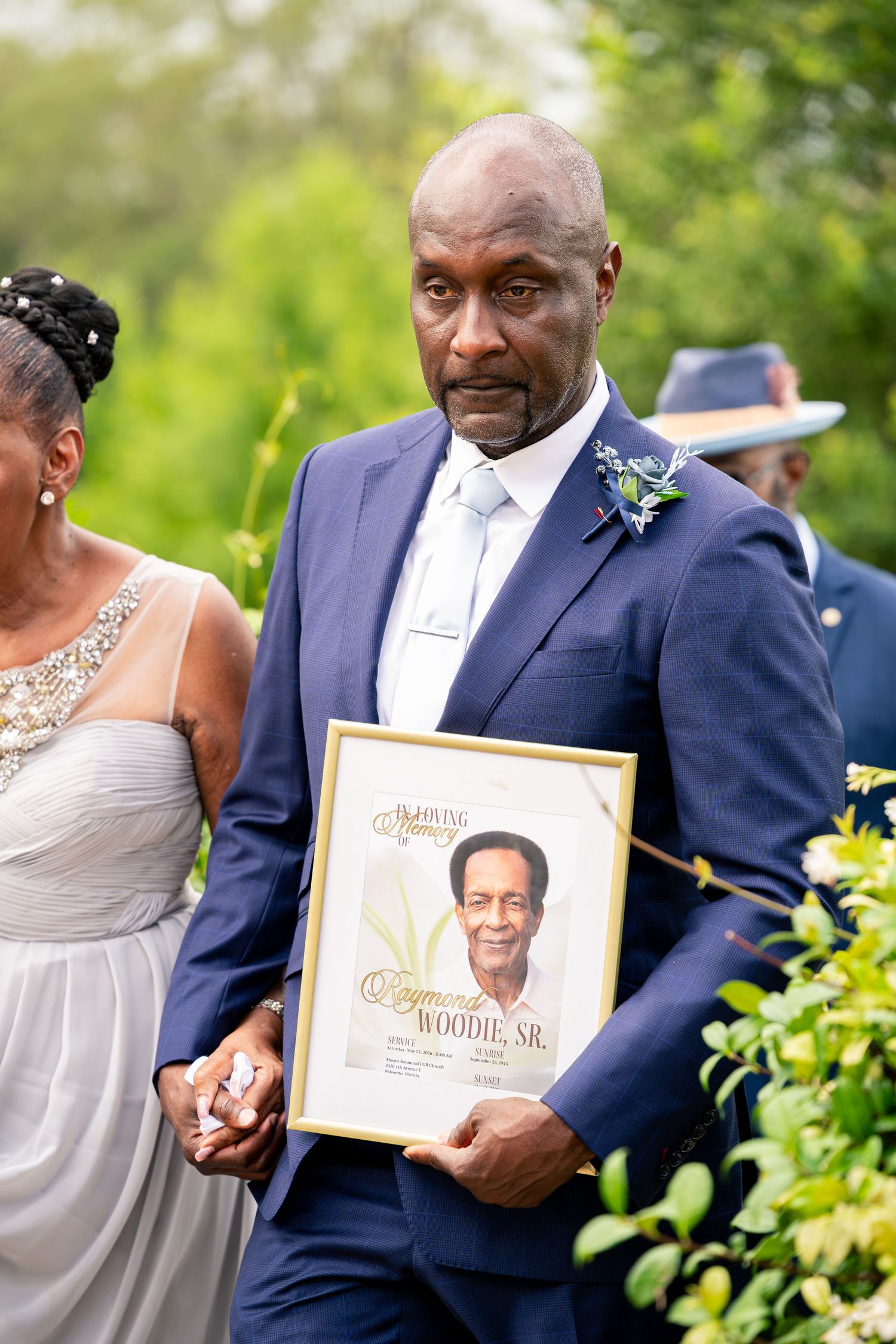 Man in navy suit holds memorial portrait of Raymond Woodie Sr. at wedding ceremony outdoors.