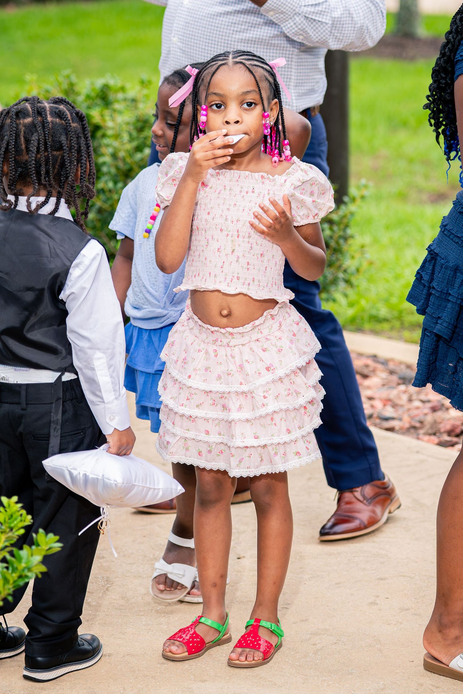 Adorable young flower girl in pink ruffled dress with colorful earrings stands at outdoor wedding ceremony.