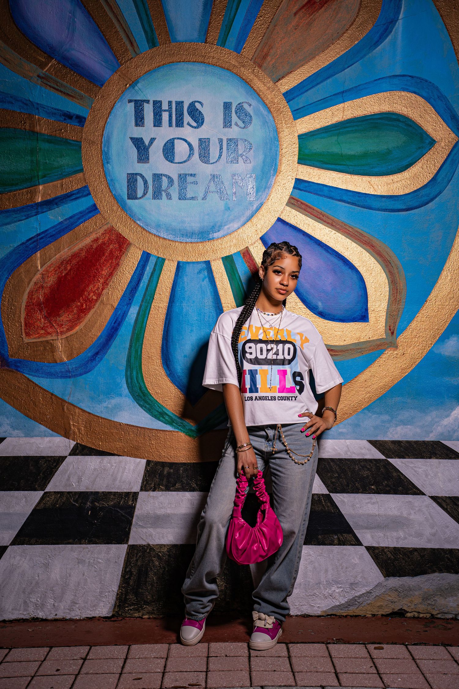 Young woman in 90210 Hills graphic tee and jeans standing before vibrant floral dream mural.