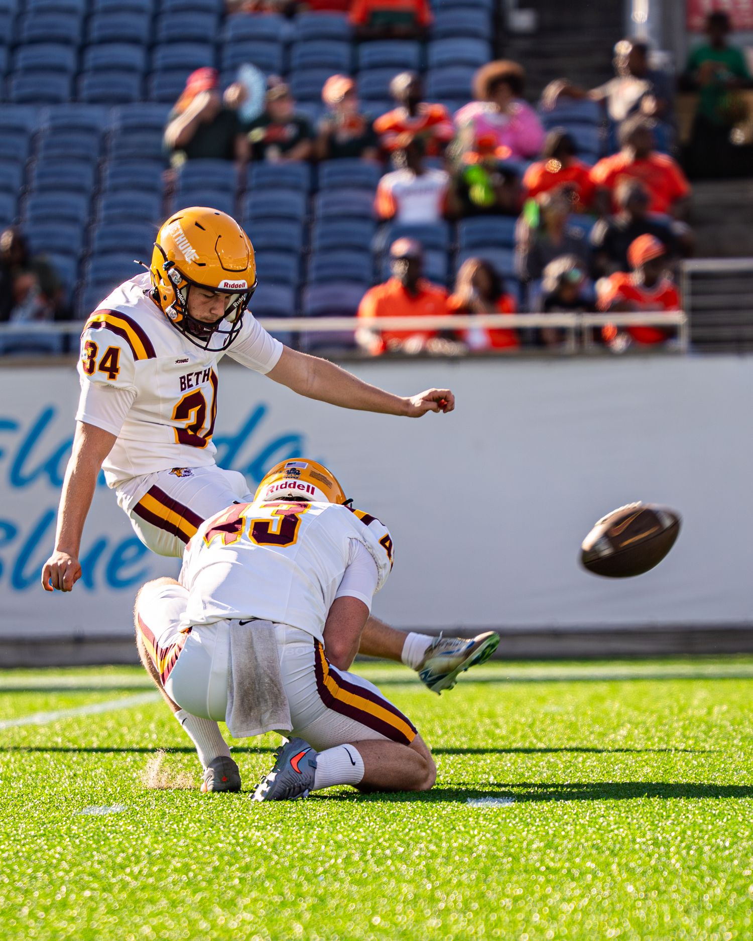 Football kicker #34 in gold helmet executes field goal attempt with holder assist during game.