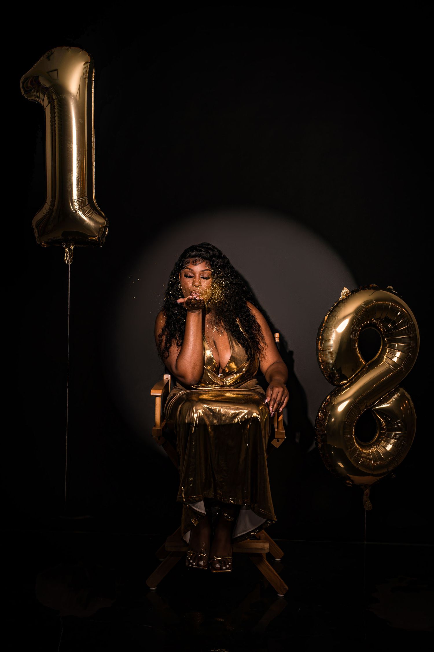 Woman in gold dress sitting between number 18 gold balloons against black background.