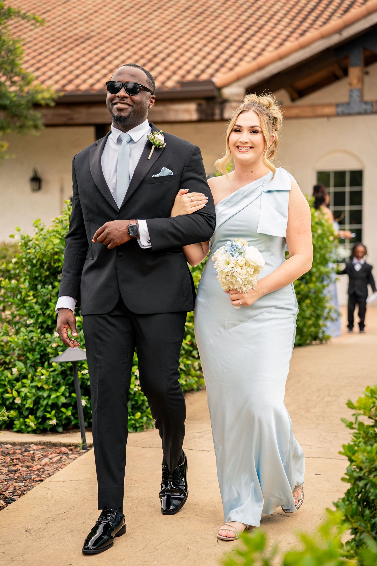Groomsman in black suit escorts smiling bridesmaid in light blue gown at outdoor wedding ceremony.