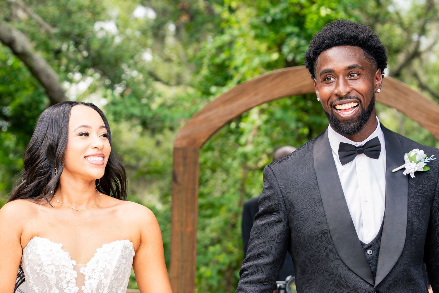 Happy bride and groom smile together under wooden arch at outdoor wedding venue.
