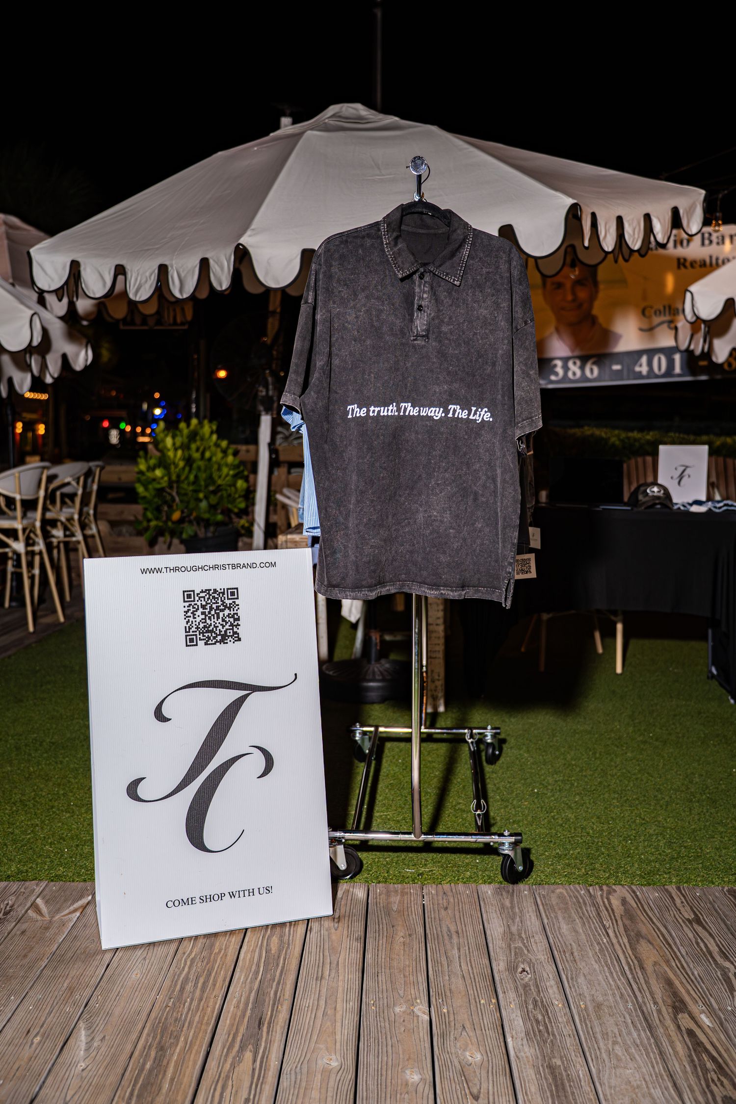 Dark gray acid wash polo shirt with white text on a rack beside a TC brand sign at an outdoor market.
