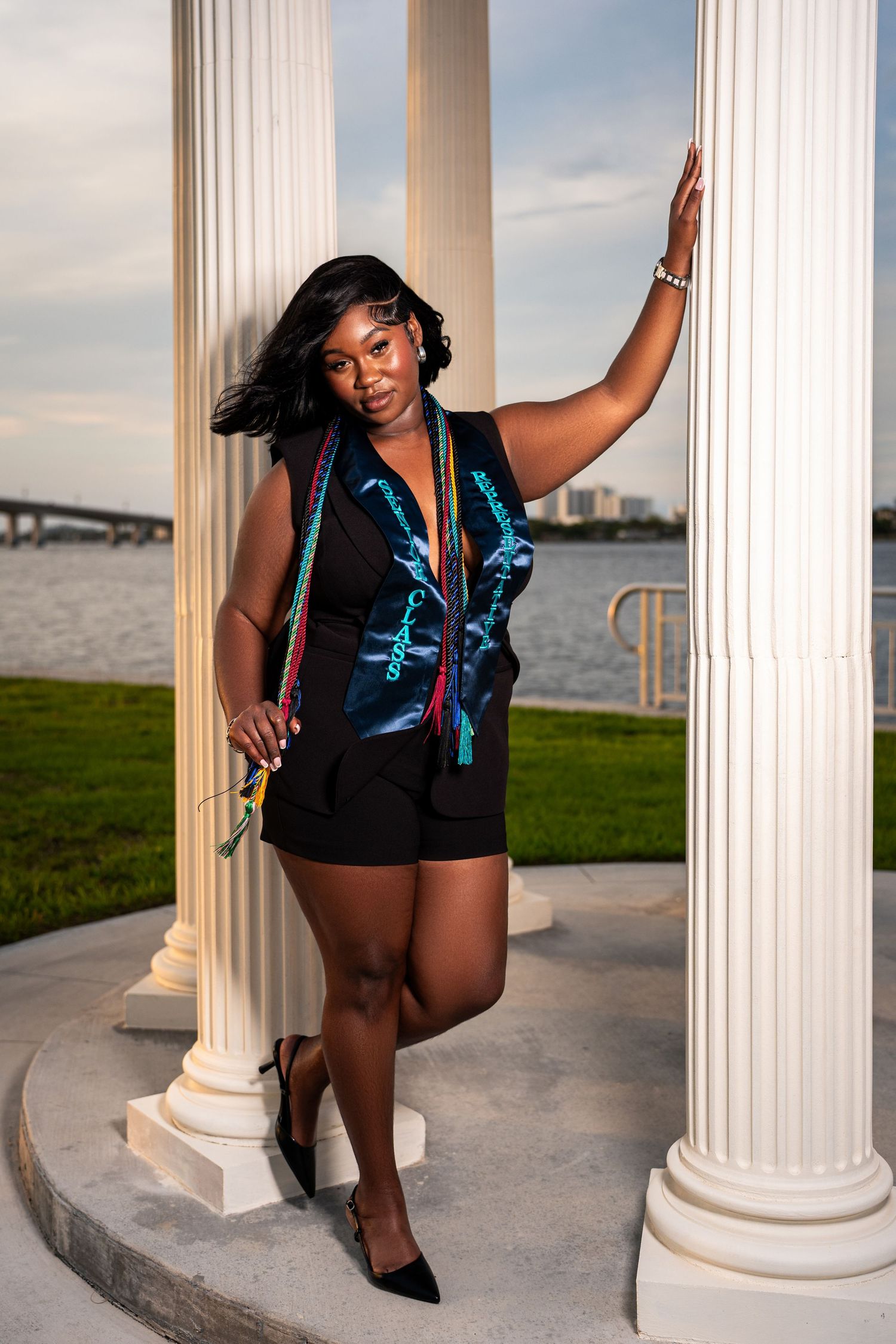 Black female graduate in black outfit with colorful graduation stole posing between white columns near waterfront.