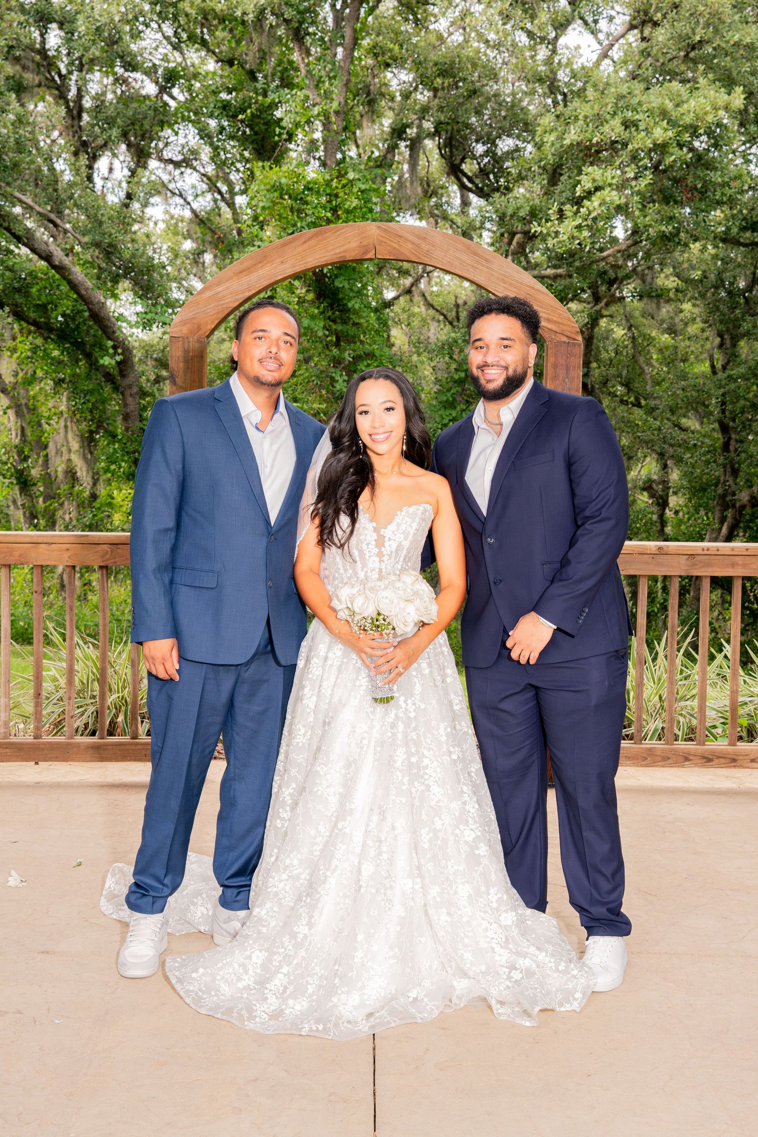 Bride in lace gown poses with two men in navy suits under wooden arch.