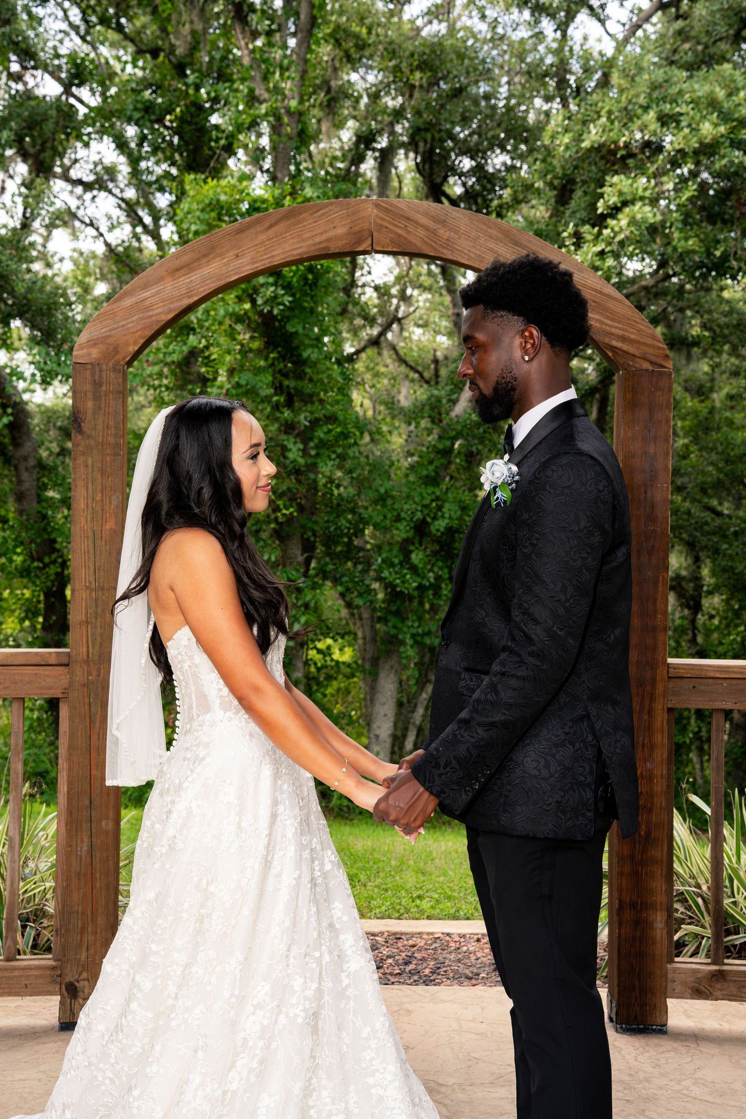 Bride and groom hold hands gazing at each other under rustic wooden arch surrounded by lush greenery.