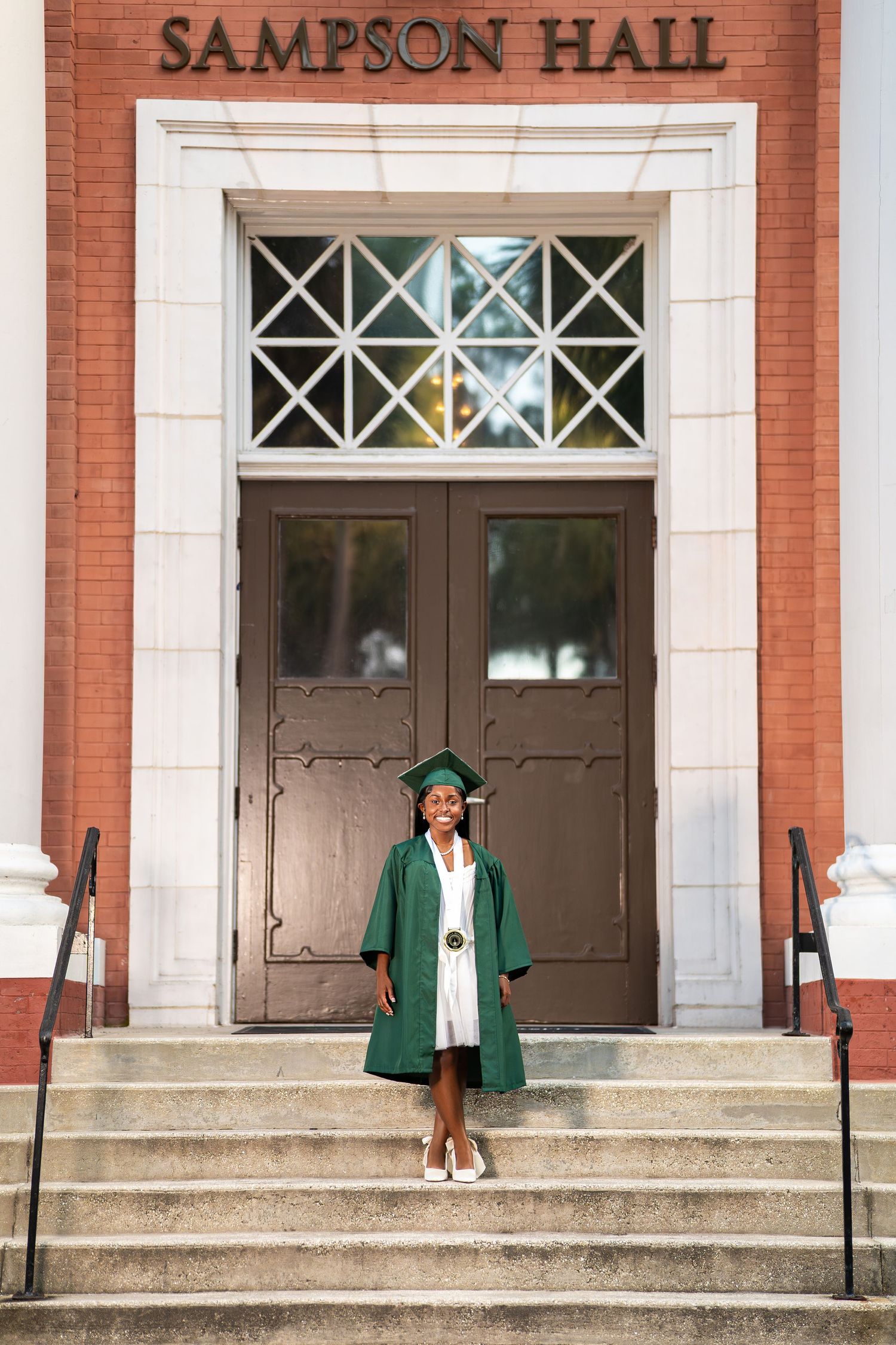 Graduate in green cap and gown stands proudly on steps before Sampson Hall brick building entrance.