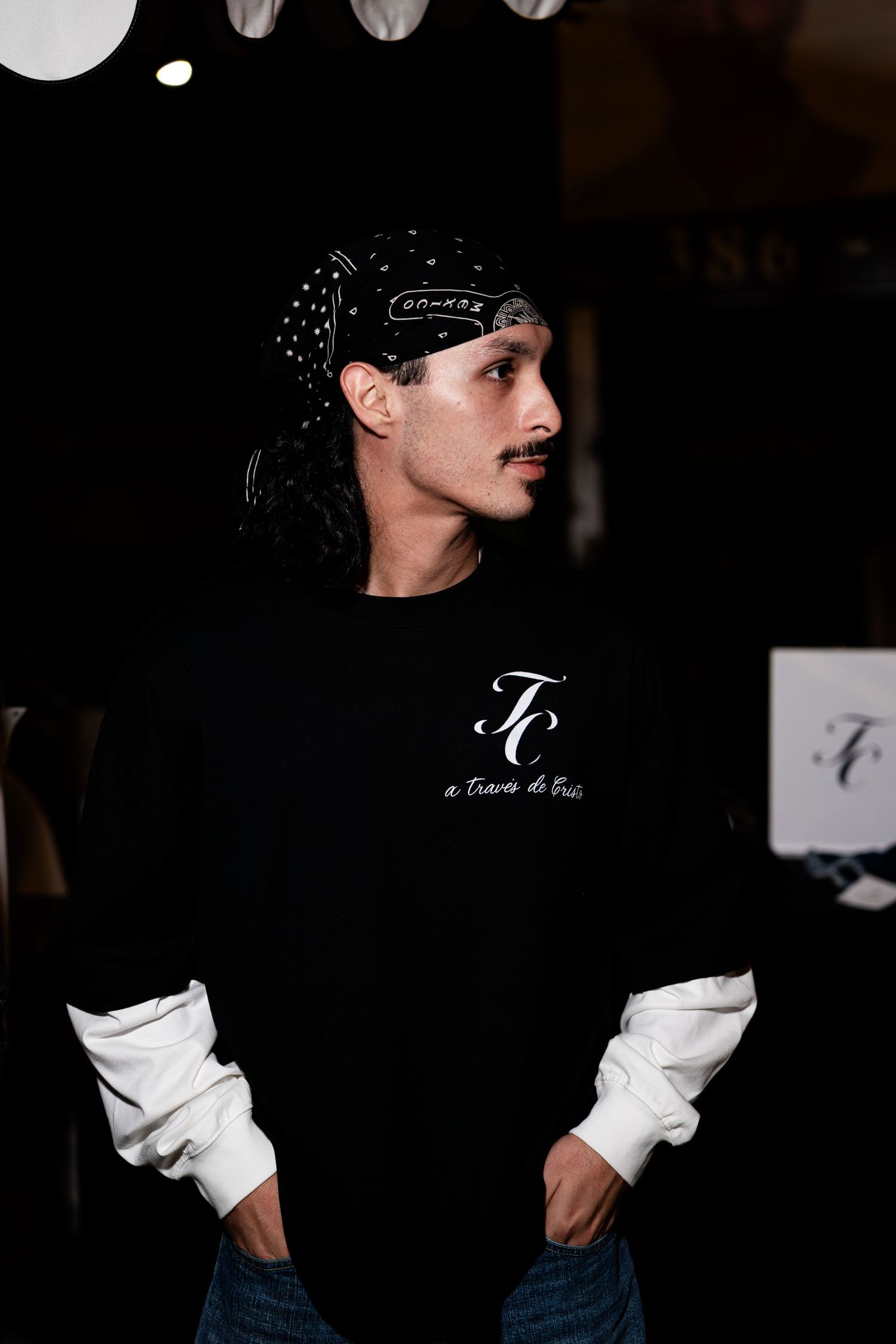 Man in black branded crewneck sweatshirt and bandana posing at nighttime outdoor clothing event.