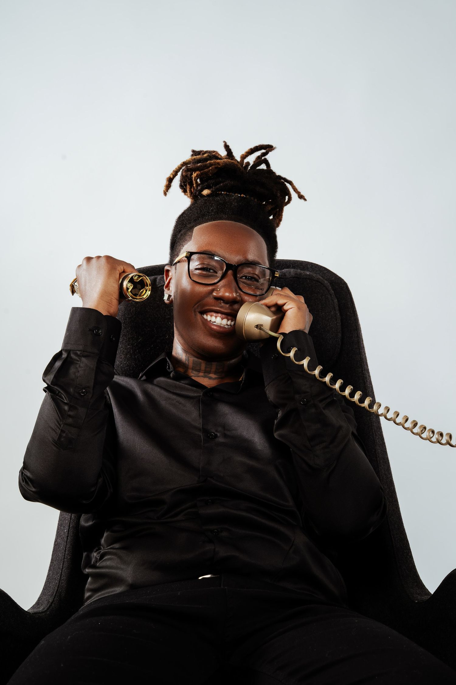 Smiling young Black man with dreadlocks holding a gold vintage telephone while seated in a dark chair.