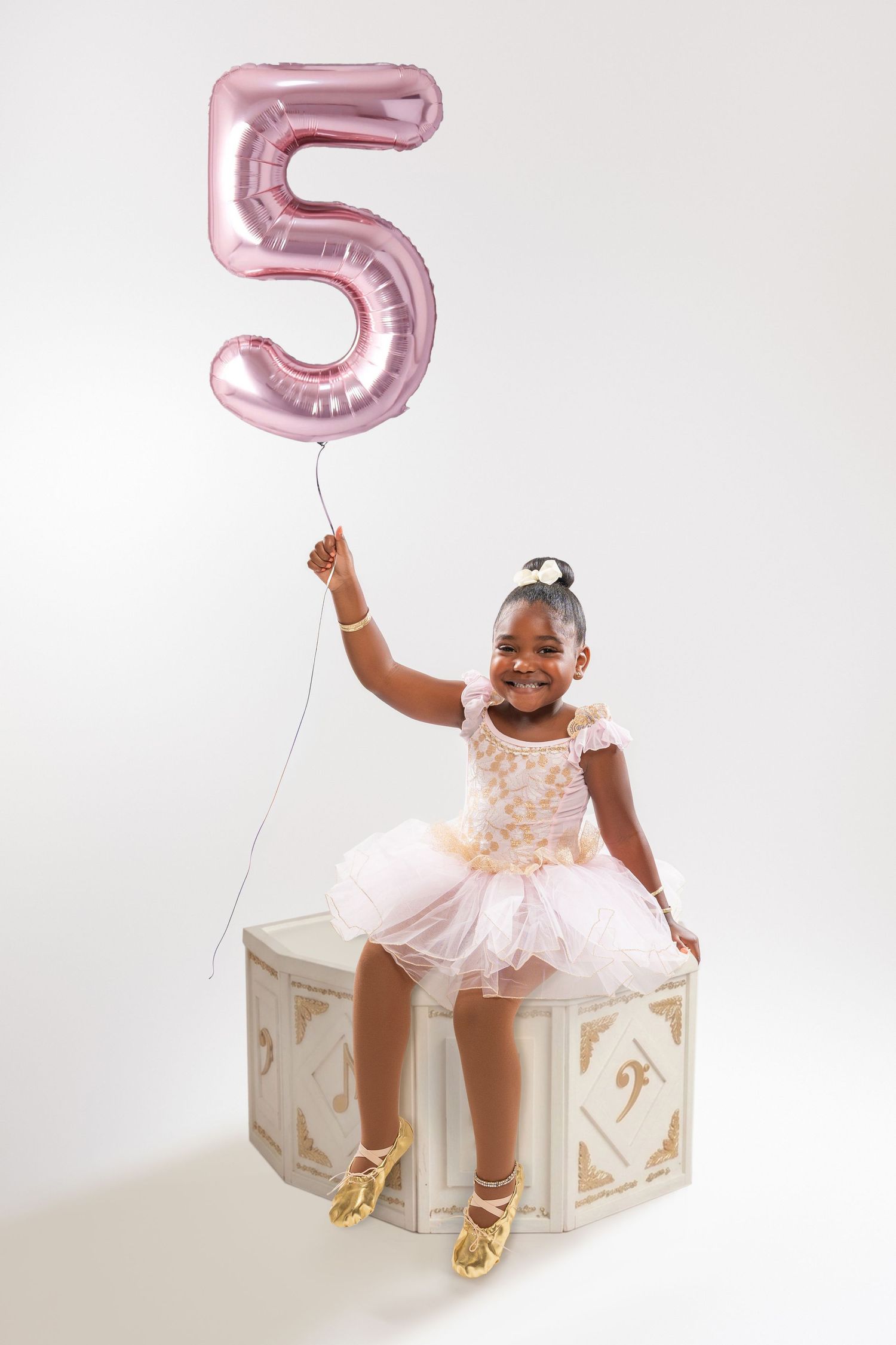 A smiling girl in a pink tutu sits on a decorative box, holding a pink number 5 balloon, celebrating her 5th birthday.