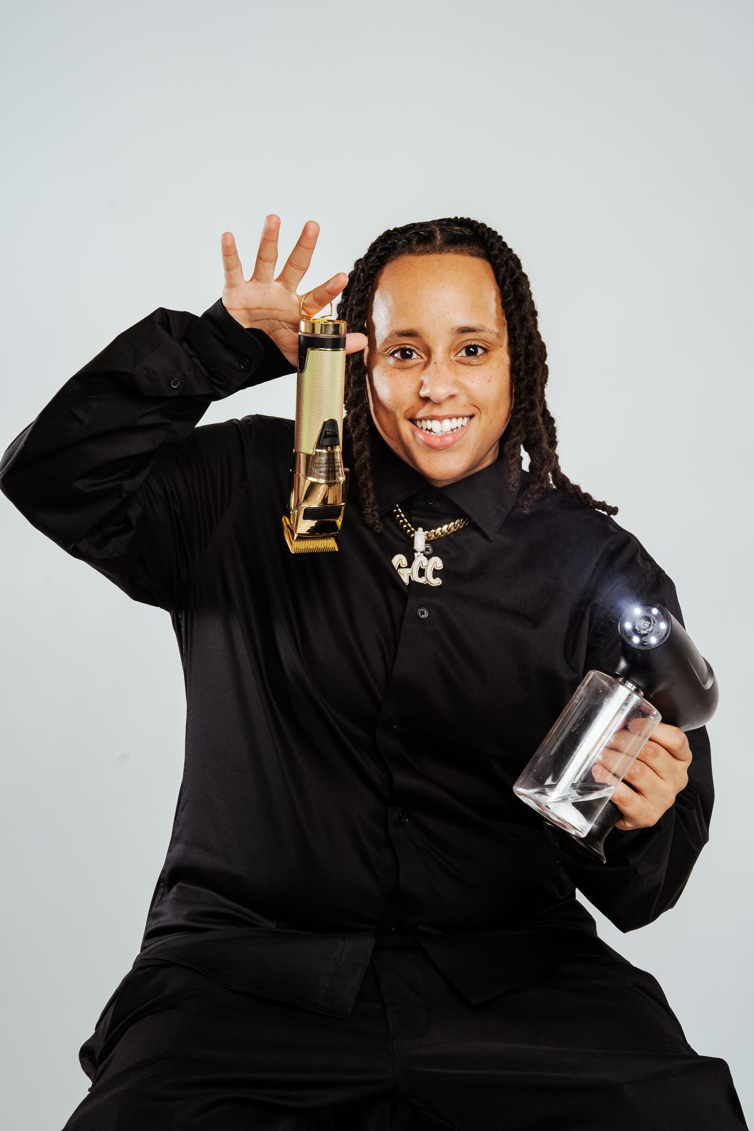 Smiling person in black outfit holding gold bottle and glass cup with chain necklace.