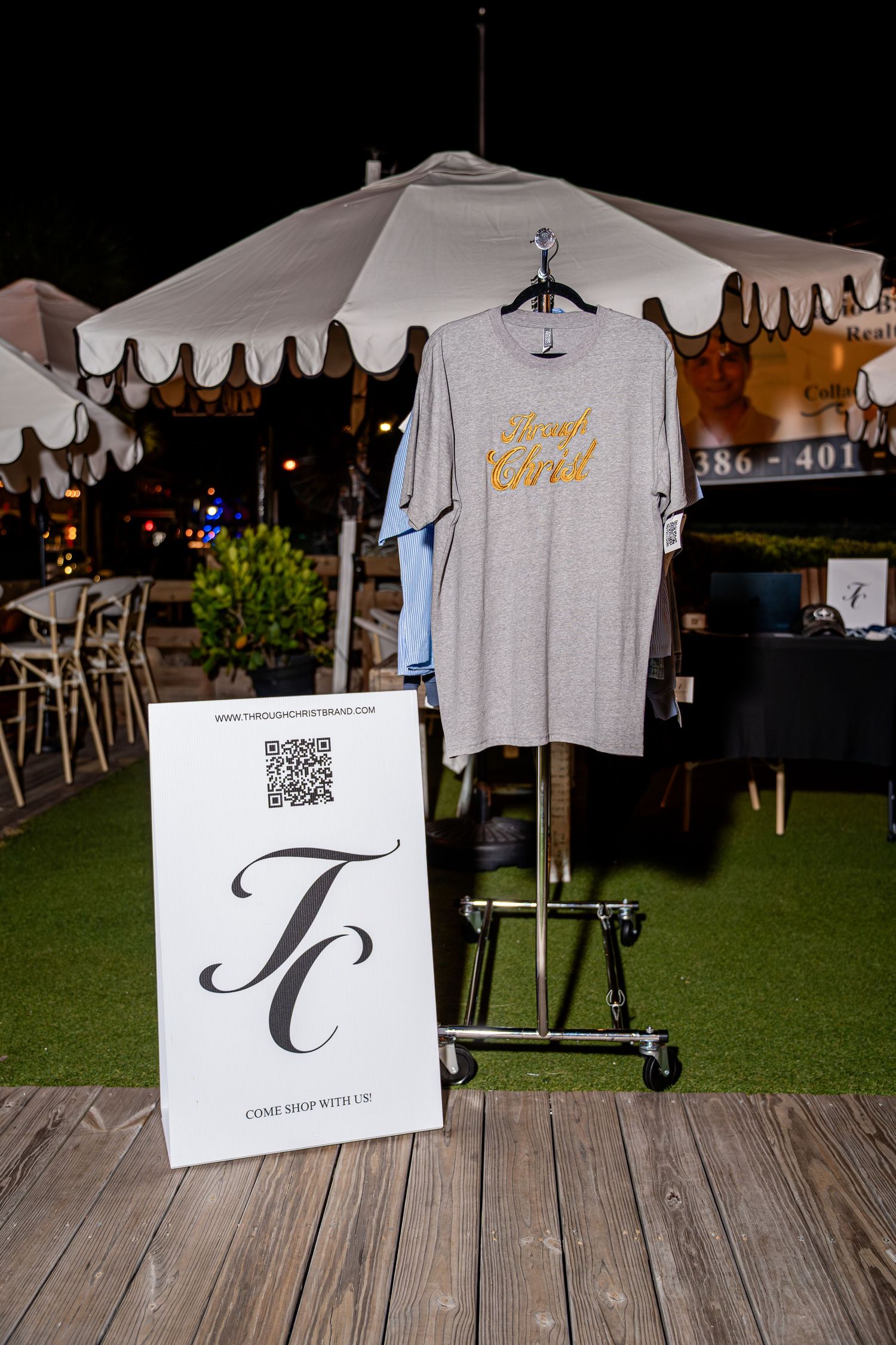 Gray t-shirt with gold Through Christ graphic on a rack beside a TC brand sign at an outdoor event.
