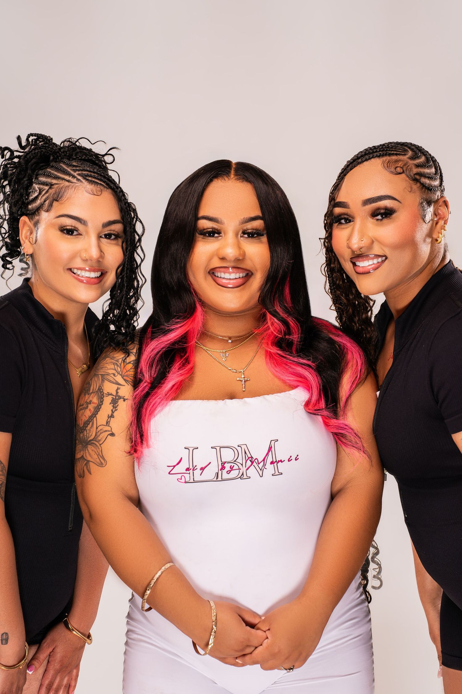Three women pose together, center wearing white LBM top with pink hair highlights, flanked by two women in black outfits.