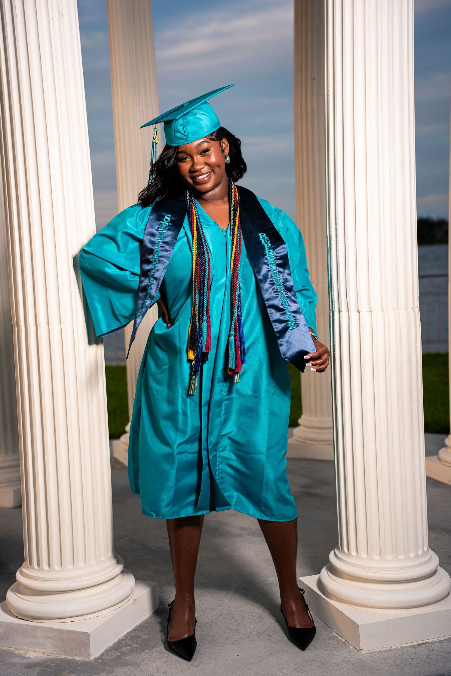 Smiling graduate in teal cap and gown with colorful honor cords poses between white columns outdoors.