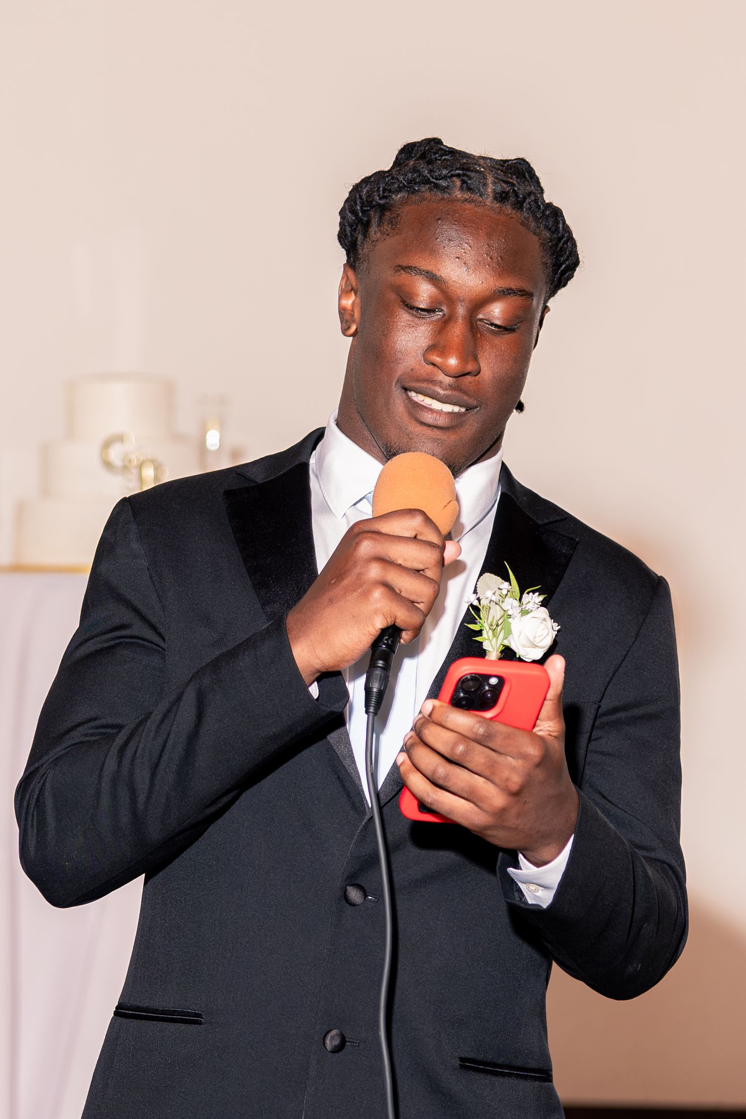 Young man in black tuxedo delivers heartfelt wedding speech while reading from his phone at reception.