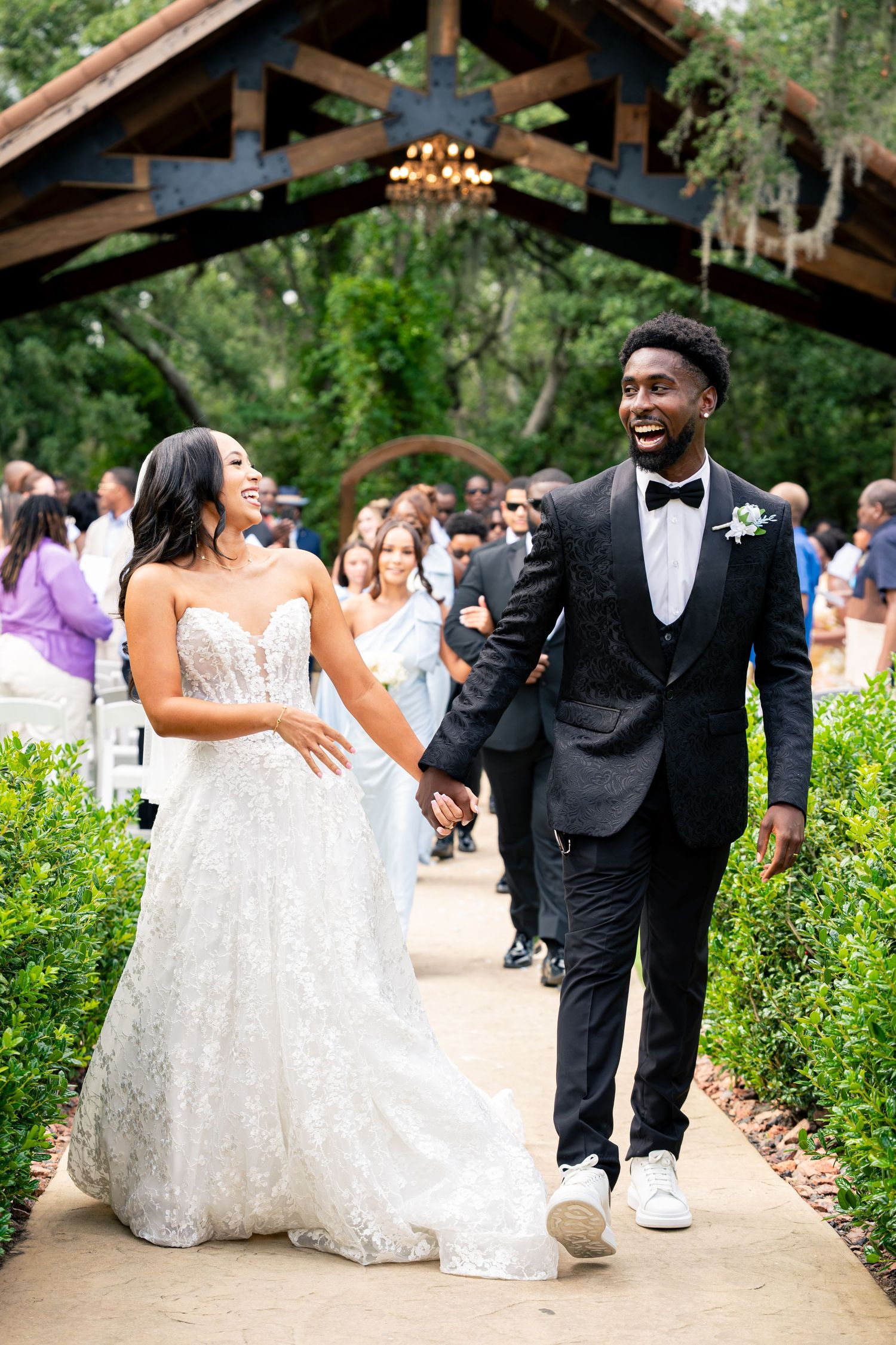 Newlyweds walk hand in hand down garden aisle, bride in lace gown and groom in black tuxedo, both laughing.