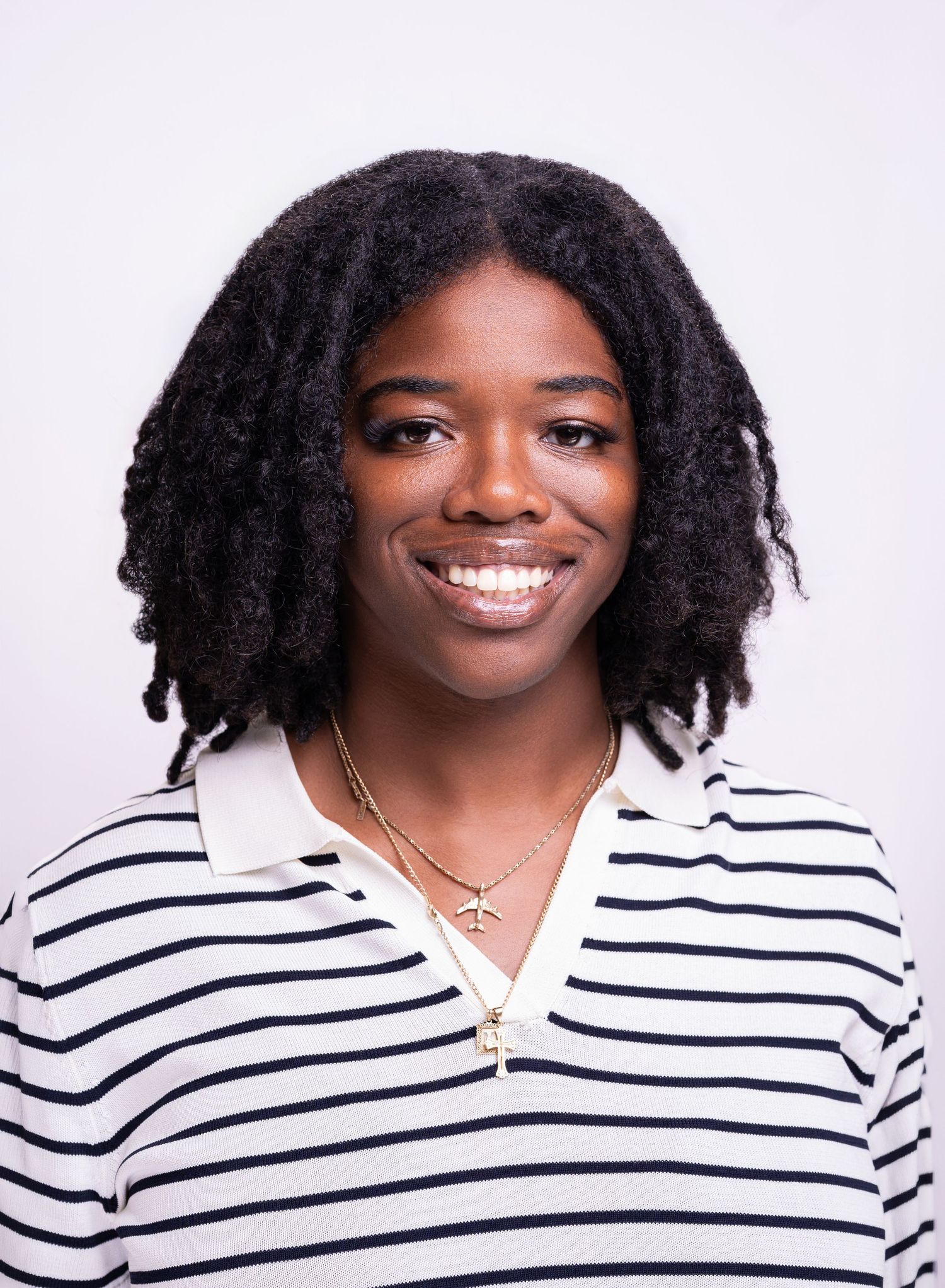 Young Black woman with natural hair wearing a striped polo shirt and gold necklace, smiling against white background.