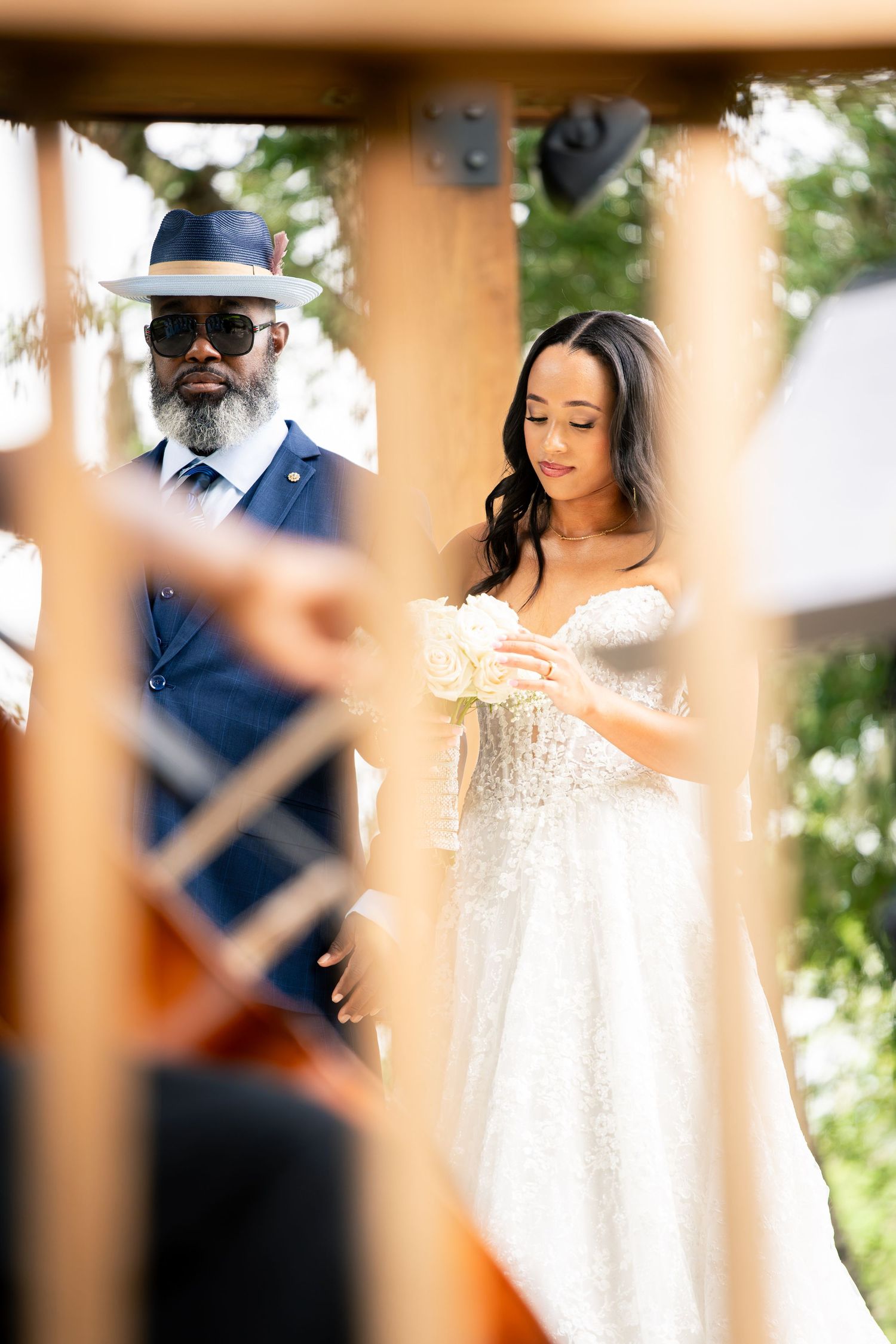 Bride in sparkly strapless gown holds white bouquet beside man in blue suit and hat at outdoor wedding ceremony.