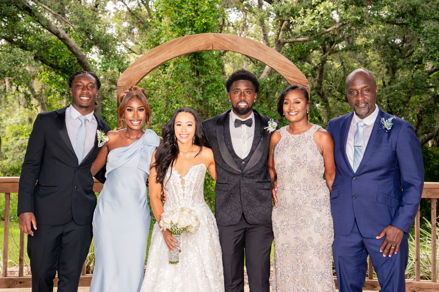 Family group portrait of six people posing under wooden arch at outdoor wedding venue.